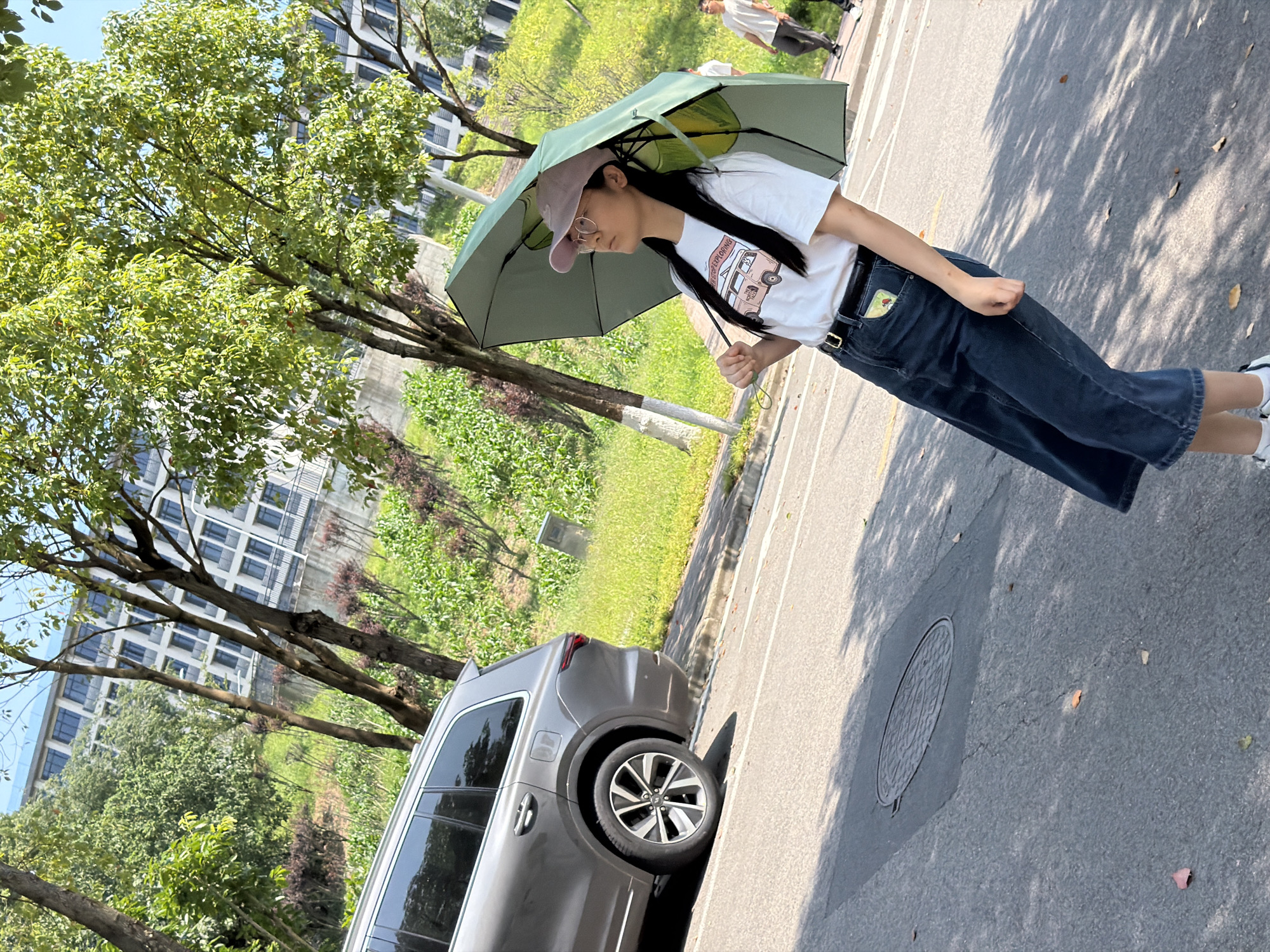 In Jiangjin, China, a young woman stands on a paved road, holding a green umbrella to shield herself from the sun. She wears a white t-shirt with a graphic print and a dark denim skirt, accessorized with a black belt. Her pink baseball cap is tilted forward, and she is wearing glasses. To her left, a grey SUV is parked parallel to the road. The background features a grassy slope with lush green trees and a modern building with multiple floors and windows. The scene is illuminated by bright sunlight, casting shadows on the road. Another person, a male wearing a white t-shirt and grey pants, is visible walking in the background to the right.