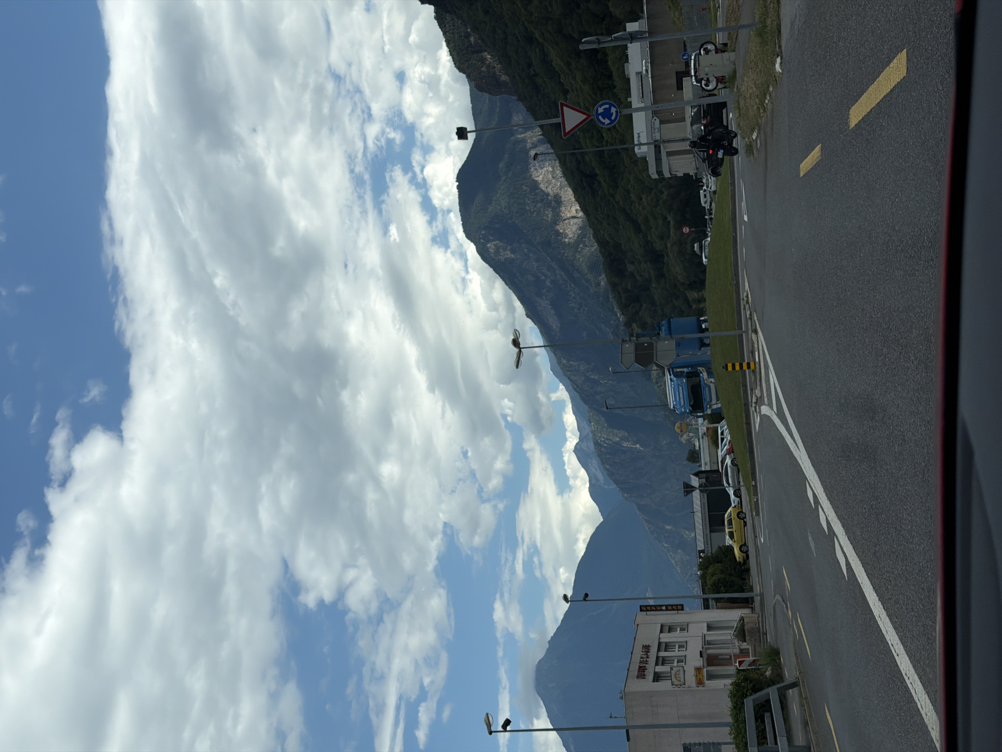 This media file captures a scenic view of a road with mountains in the background, likely taken from within a vehicle. The scene is dominated by dramatic clouds and a clear blue sky.
The road, paved with asphalt, features white lane markings and yellow dashed lines, suggesting it is a well-maintained road, possibly a highway or a major route. Several vehicles are visible on the road and in a parking area to the side. These include trucks, cars, and a motorcycle. A yellow car is parked near a building on the right side of the road. A blue truck is also visible.
The mountainous landscape is a prominent feature, with steep slopes covered in green vegetation. The scale of the mountains is emphasized by the vastness of the sky and clouds above.
Streetlights and traffic signs are also present, including a roundabouts sign and a triangular warning sign. A building with multiple windows and signage is visible on the right side of the road. Text is visible on this building, but it is difficult to read clearly.
The time of day appears to be daytime, indicated by the bright sunlight and the presence of shadows. The weather seems pleasant, with a mix of sun and clouds. The overall impression is one of a picturesque journey through a mountainous region. The location context provided is La Balmaz, Switzerland.