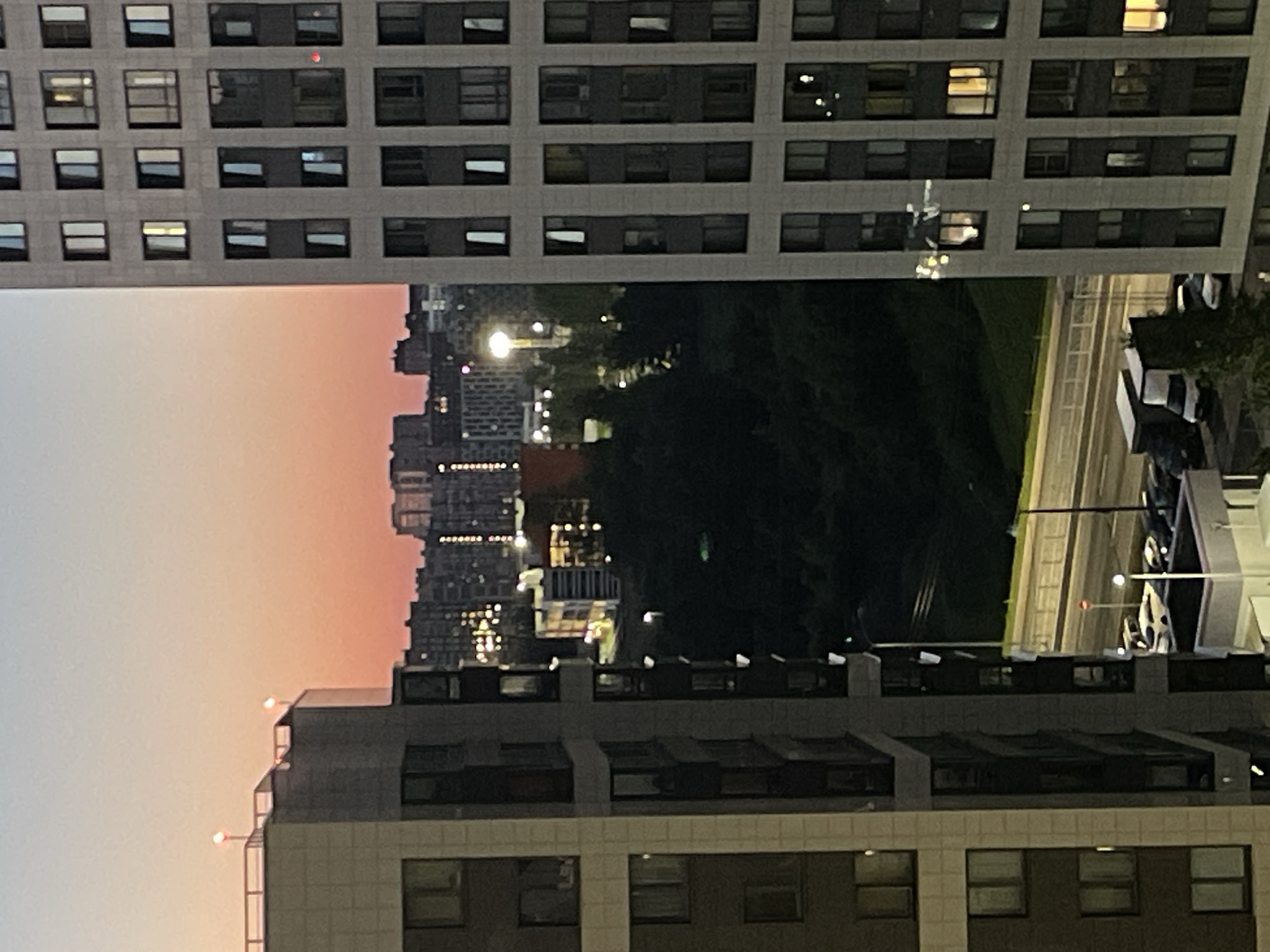 The image depicts a dusk scene in the city of Sinyavino, Russia, viewed from a high vantage point. In the foreground, two large apartment buildings dominate the frame. The building on the left is characterized by its beige and dark gray facade, with numerous windows visible. The building on the right, taller and more prominently featured, has a light gray exterior with a grid of dark-framed windows, many of which are illuminated from within.
The background reveals a cityscape with more apartment buildings, some of which also show illuminated windows. A densely wooded area or park is visible between the foreground buildings and the distant city. A road with streetlights runs horizontally across the middle ground, with several parked vehicles, including cars and vans, visible along its edge. The sky above is a gradient of light blue and soft orange, indicative of sunset. Two red lights are visible on the rooftop of the building on the left, possibly navigational lights. A bright, solitary streetlight illuminates a section of the park area. There are no people clearly visible in the image.