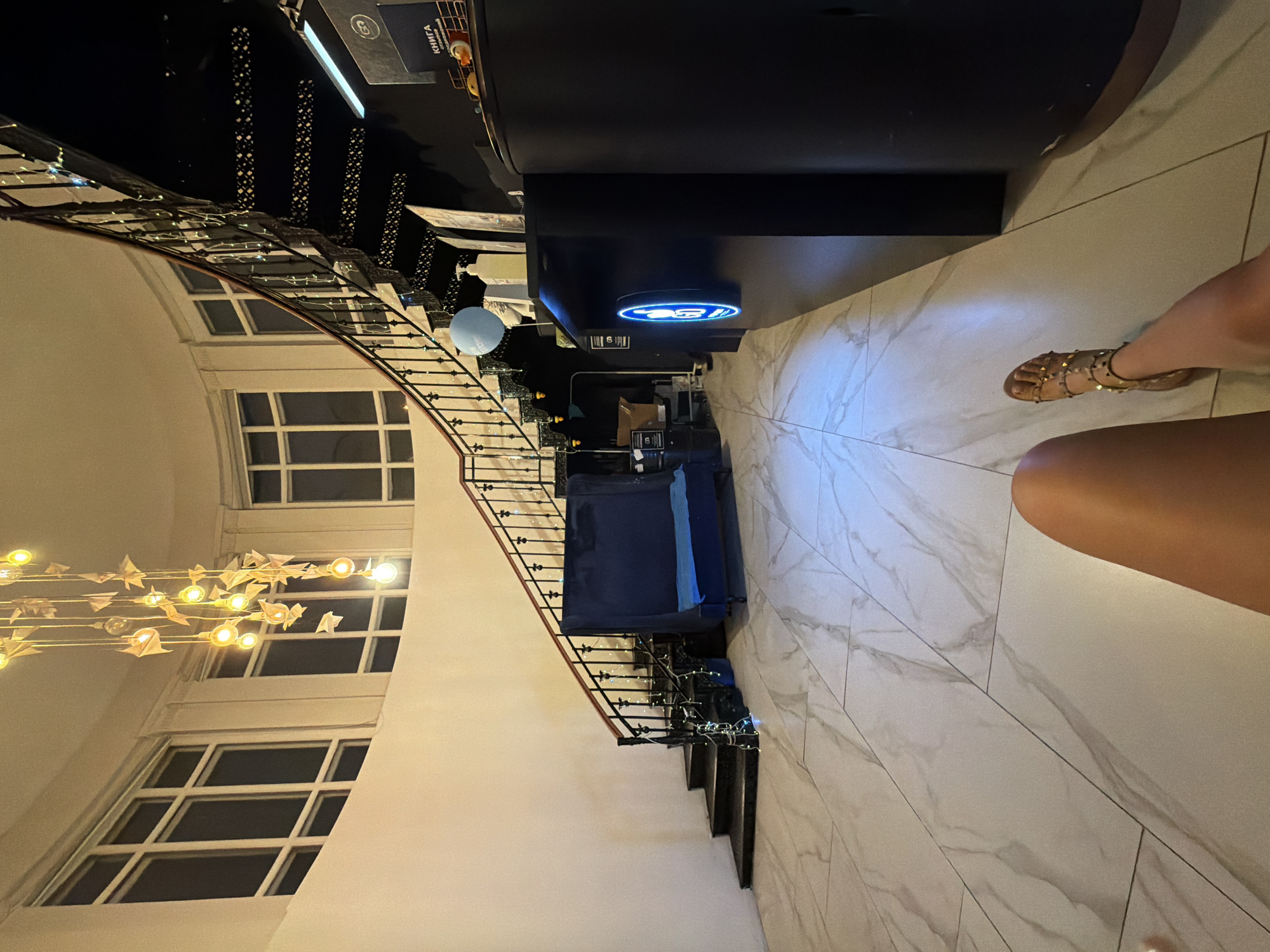 The image shows the interior of a reception area with a prominent curved staircase. The staircase has a dark metal railing and is decorated with string lights. Above the staircase, a unique chandelier made of light bulbs and paper airplanes hangs from the ceiling. The walls are painted a light cream color, and large windows with multiple panes are visible on the upper level. The floor is covered in large, light-colored marble-patterned tiles.
To the right of the staircase, there is a black reception desk with a blue illuminated logo on its front. A light blue balloon is attached to a stand on the desk, along with a hand sanitizer dispenser. Various items are placed on the desk, including what appears to be a book with Russian text.
In the foreground, a person's legs and feet are visible, wearing gold studded sandals. The person appears to be sitting or reclining on the floor.
The image is set in Moscow, Russia, specifically at the reception of the "Artplay Design Center" (Центр дизайна Artplay).