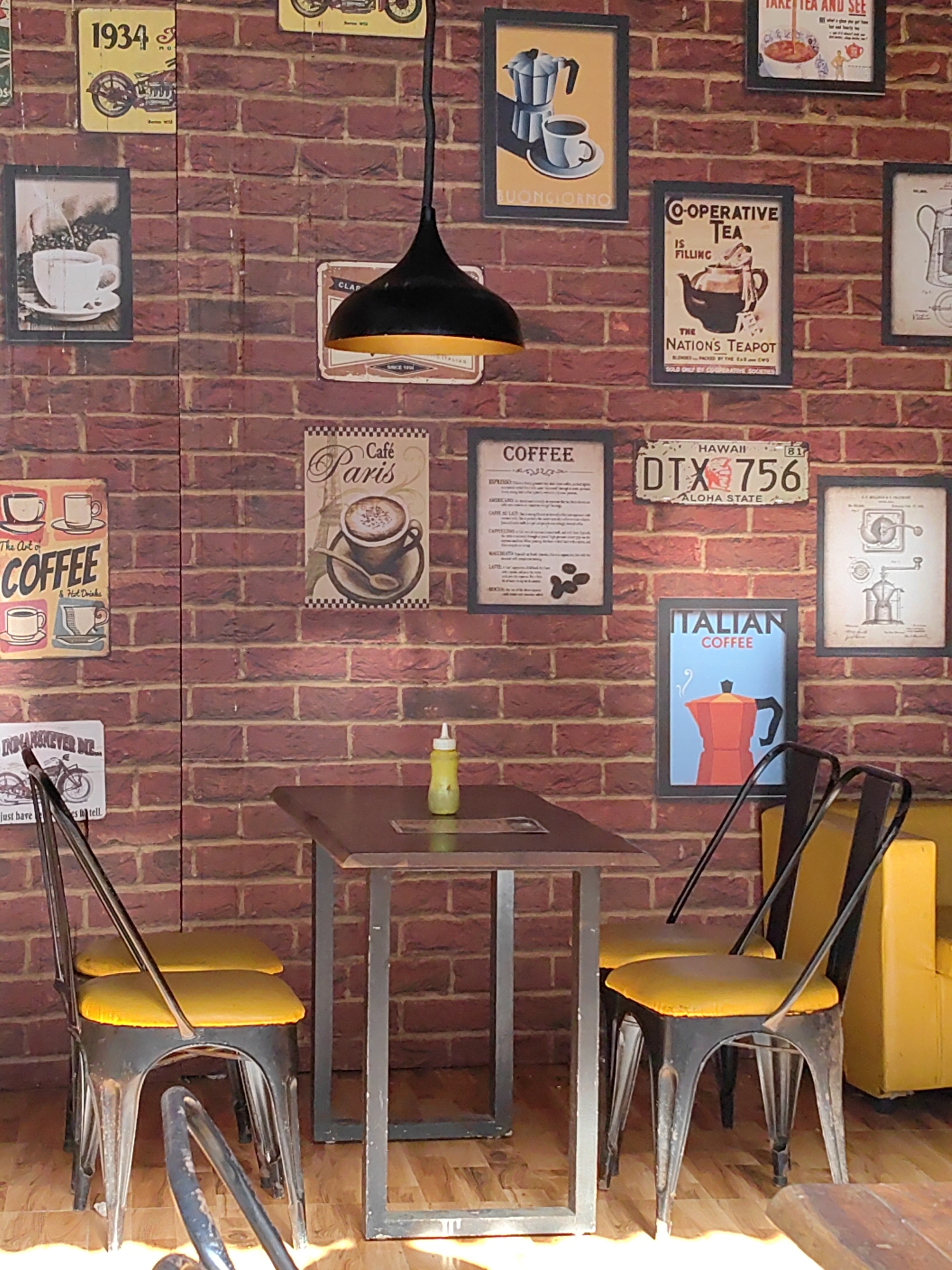 This is an eye-level, medium shot of a cafe wall with various posters and a small table with two chairs. The wall is a brick pattern, and on it are several framed posters related to coffee and tea. The posters include a "1934" motorcycle, "Cafe Paris", a coffee menu, a "Co-operative Tea" poster, a "Hawai DTX 756 Aloha State" license plate, a "Italian Coffee" poster, and a poster with a coffee grinder. A black hanging lamp with a yellow interior is visible at the top center of the image. In the foreground, a dark brown table with a metal frame sits between two metal chairs with yellow cushions. A yellow squeeze bottle is on the table. The floor is made of wood. The cafe is located in an unknown city, in an unknown location.
