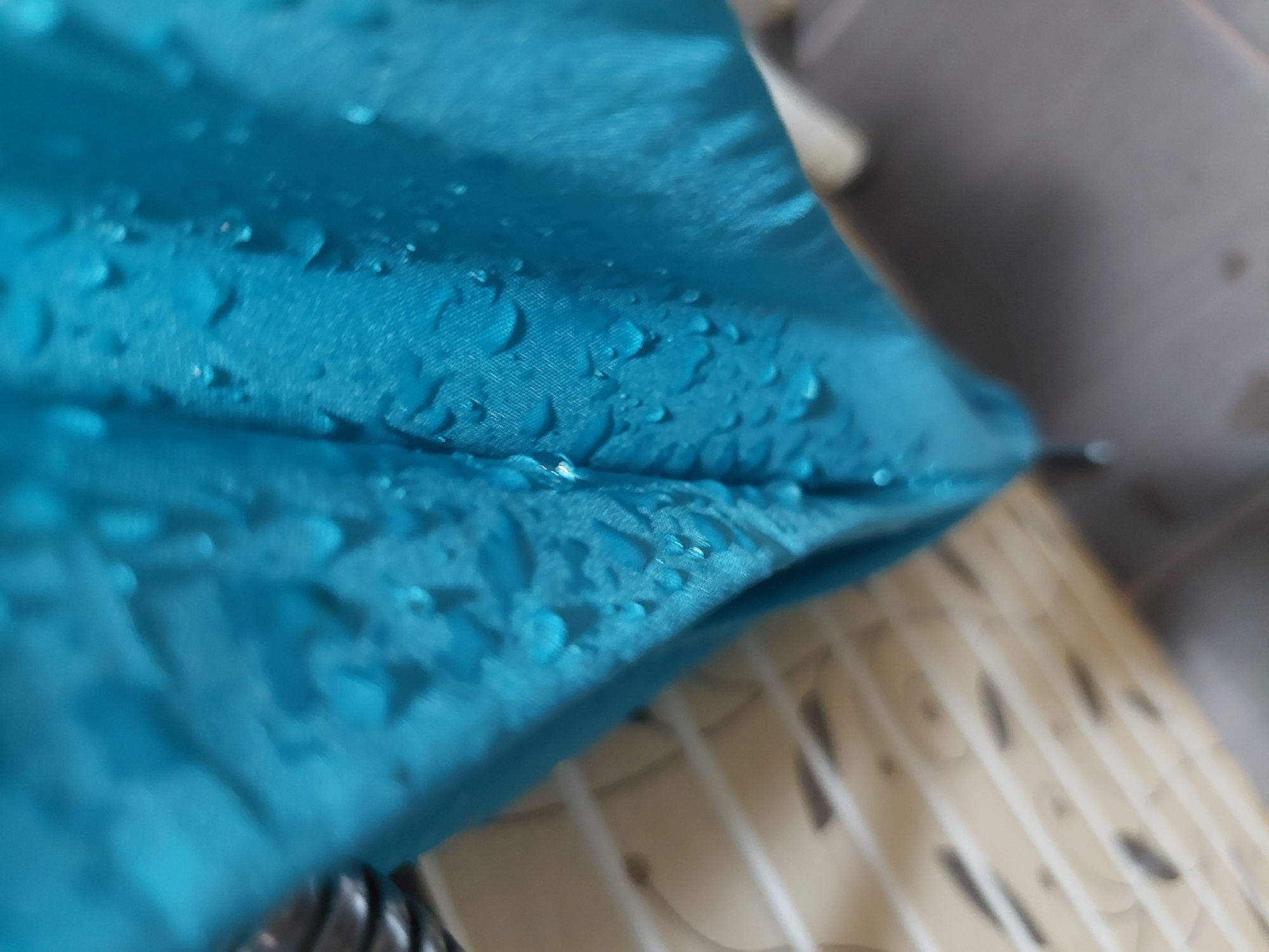A close-up shot focuses on a section of wet, turquoise fabric, possibly an umbrella or tent, covered with numerous water droplets. The fabric texture is visible. The background is blurred, revealing what appears to be a tiled floor or wall with a pattern of beige and dark brown. A hint of a white or light-colored object is seen near the top edge. No people are visible. The image suggests an indoor setting after or during rainfall. There's no information to determine whether the image was taken in Plovdiv, Bulgaria, or elsewhere.