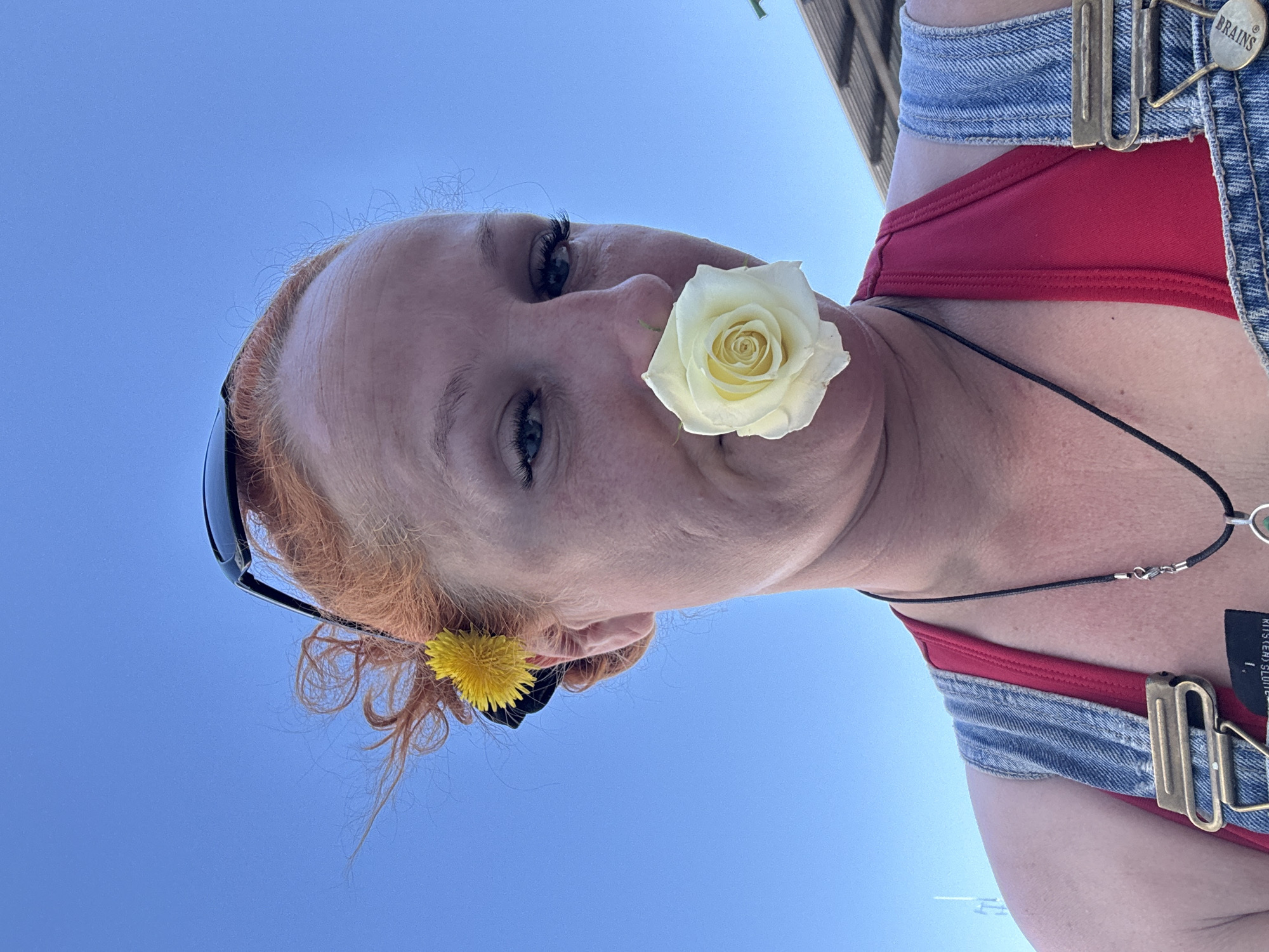 The image depicts a person with red hair, sunglasses perched on their head, and a white rose held between their nose and cheek. A yellow dandelion is tucked behind their ear. They are wearing a red top and blue denim overalls with a "BRAINS" button. A black cord necklace with a silver pendant is visible around their neck. The background shows a clear blue sky. In the far distance there is a suggestion of a building. The location is implied as Chaikovsky, Russia. No specific details in the image confirm this location.