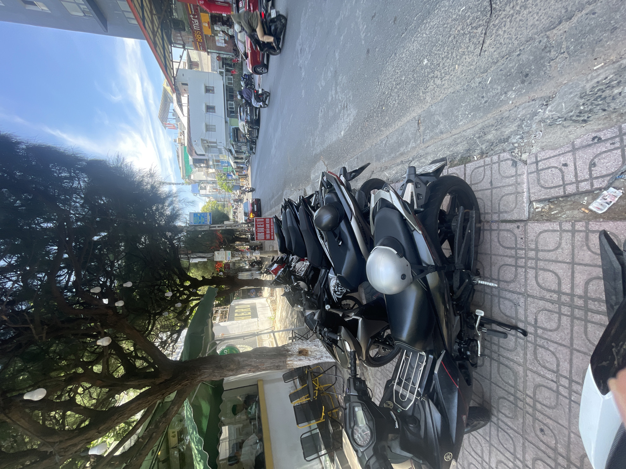 This image depicts a street scene in Bac Nha Trang Ward, Vietnam, on a bright, sunny day with scattered white clouds. The primary subjects are a row of parked motorcycles lined up along the sidewalk. The motorcycles are mostly dark-colored and appear to be scooters or smaller motorbikes, common in this region. One motorcycle in the foreground has a silver helmet resting on its seat, and another has a metal rack attached to its rear.
The setting is an urban street with buildings on one side and a sidewalk and road on the other. The buildings are a mix of modern and older structures, with awnings and some signs visible. A large, leafy tree with white globes hanging from its branches stands on the left side of the image, casting shadows onto the sidewalk. The road is paved and appears to be a typical street in a populated area, with some debris and cigarette butts on the pavement.
In the background, more motorcycles and some cars are visible on the street, suggesting moderate traffic. The overall atmosphere is that of a typical day in a bustling Vietnamese city, with the bright sunlight indicating it is likely daytime, possibly late morning or early afternoon. There are no people prominently featured in the immediate foreground, but some figures can be seen in the distance on the street and sidewalks. The image's orientation is vertical, with the sky and distant buildings at the top and the motorcycles and sidewalk in the foreground. No specific event or activity is clearly taking place beyond the general scene of parked vehicles and the urban environment.