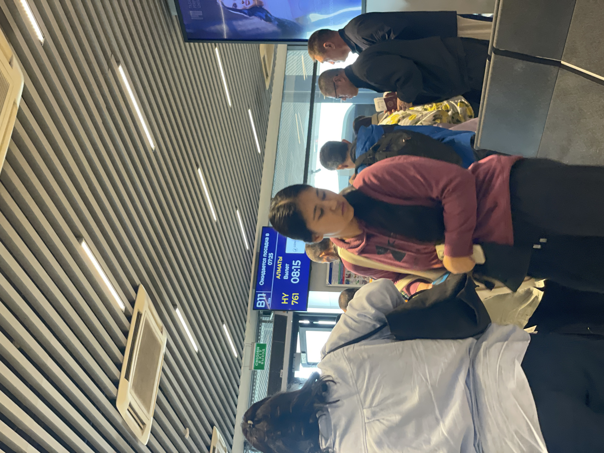 The image shows people at an airport gate in Tashkent, Uzbekistan. Passengers are waiting in line near gate B11. A flight information display shows "Almaty" as the destination, flight HY761, scheduled to depart at 08:15, with boarding expected at 07:25. People are dressed in casual clothing.
