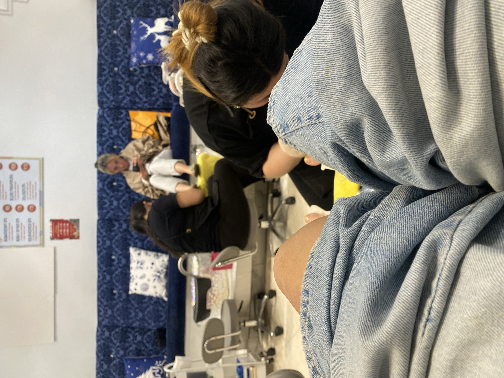 The image shows the interior of a nail salon. In the foreground, a person wearing a denim jacket is seated. To their left, a nail technician with a bun is working on someone's nails. Further back, another customer is sitting on a blue patterned sofa, using a phone. There are chairs, tables, and decorative pillows. The location is the city of New York, United States of America. There are signs posted on the wall.