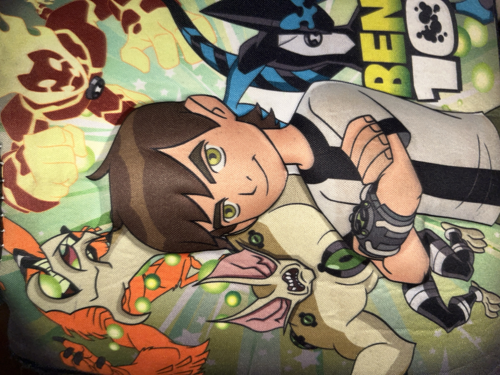 The image shows a close-up of a patterned fabric, likely from a backpack or piece of clothing, featuring characters from the Ben 10 animated series. The main subject is Ben Tennyson, depicted in his classic outfit with brown hair and green eyes, arms crossed. Behind him, various aliens from the show are illustrated: a creature resembling an orange fox with sharp teeth and claws on the left, a being made of fire on the top right, and a bat-like alien with large ears and sharp teeth on the left, behind Ben. To the right of Ben, a black and blue alien with a stylized design is visible. The background is a light green with subtle star and bubble patterns. Text that reads "BEN 10" is prominently displayed in yellow and white letters on the lower right side. The fabric appears to be textured, indicating a textile material. The image is illuminated from above and to the left, casting faint shadows. There is no indication of the city of Gura Top, Nigeria in the image itself.