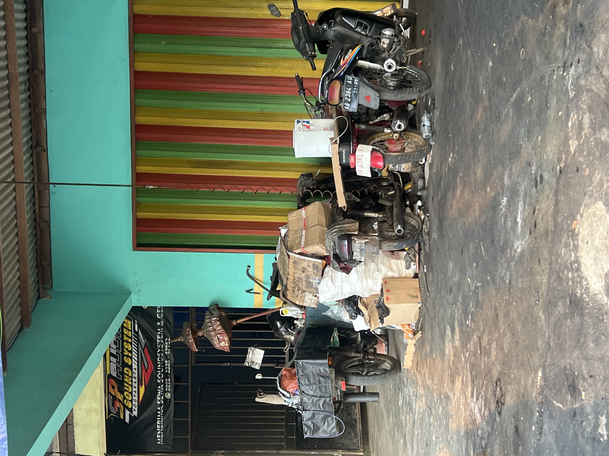 The image depicts a cluttered outdoor scene in Tlogoadi, Indonesia, likely in front of a business. In the foreground, the ground is a dark, stained concrete surface, with some lighter debris and oil stains.
Dominating the mid-ground are several motorcycles parked closely together, some appearing to be loaded with various items. One motorcycle on the left has a black cargo box attached to the rear, filled with bags and what appears to be a tall, decorative umbrella-like object with a dark red, ornate top and a slender pole. Another motorcycle in the center is heavily laden with boxes and plastic-wrapped bundles. To the right, two more motorcycles are visible, one red and one black.
Behind the motorcycles, a corrugated metal wall with vertical stripes of red, yellow, and green forms the backdrop. Above this wall, a teal-colored building facade is visible, with a corrugated metal roof extending overhead. A banner hangs on the left side, advertising "SP AUDIO SOUND SYSTEM YOGYAKARTA," and includes text in Indonesian indicating rental services and phone numbers. Various objects, including bags of white material, cardboard boxes, and discarded items, are scattered around the base of the motorcycles, contributing to the sense of disarray. The overall impression is of a storage or working area, possibly related to an audio equipment rental business, with a functional but untidy presentation.