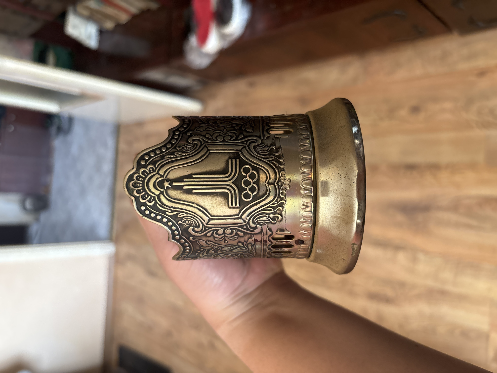 A hand holds a tarnished brass podstakannik, or Russian cup holder, with ornate detailing and a stylized Olympic rings emblem. The podstakannik is cylindrical with a flared base and a decorative, wavy rim. The background suggests an indoor setting with wooden flooring and blurred furniture, hinting at a domestic environment. The image is taken from a medium close-up perspective, focusing on the object. The location in Asenovgrad, Bulgaria is not discernible from the visual information provided.