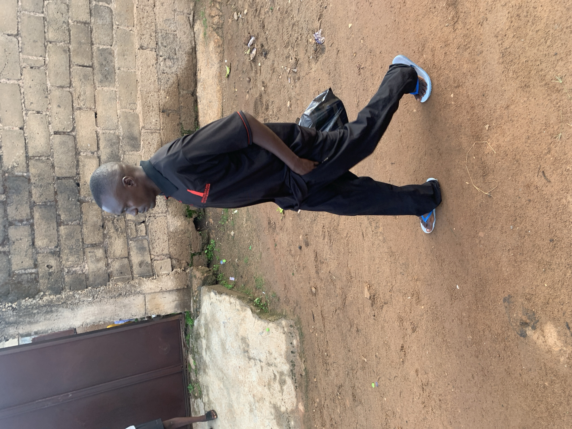 The image depicts a man walking outdoors in what appears to be a dirt or dusty area. He is wearing a black polo shirt with a red logo on the left chest and black trousers. He has a black plastic bag tucked under his right arm. He is wearing blue and white flip-flops. In the background, there is a rough brick wall and a large metal door. The ground is covered in loose dirt and some scattered debris, including what looks like small colorful plastic bottle caps and bits of paper. The scene suggests an environment that is not highly developed, possibly in a rural or semi-urban setting. The man is walking from left to right in the frame, and his body is angled away from the camera.