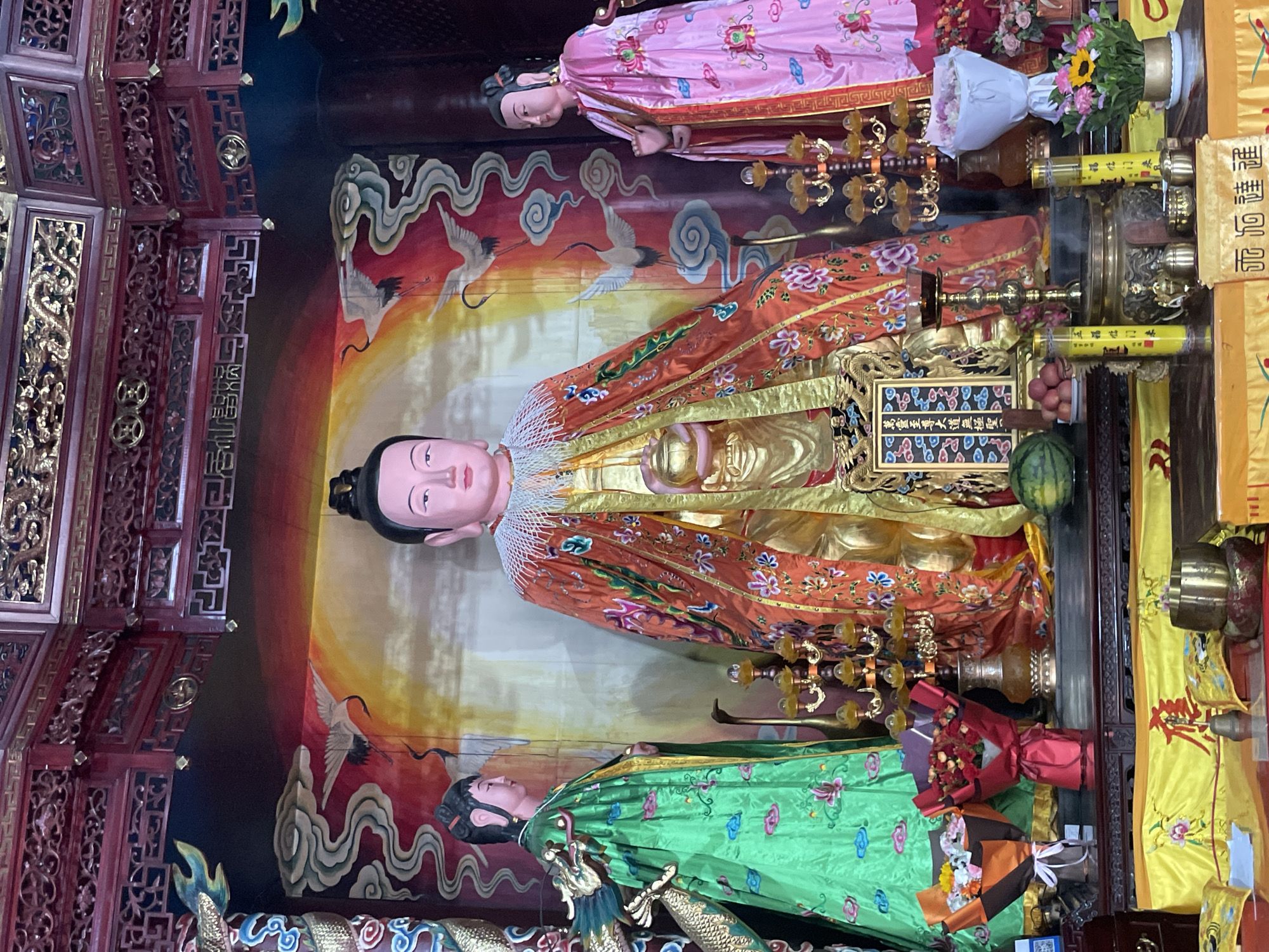The image shows a group of three Buddhist statues inside what appears to be a temple or shrine. The central figure is the largest, wearing an orange and gold robe with a serene expression. To the left is a statue in a green robe, and to the right, a statue in a pink robe. All three are positioned against a backdrop painted with cranes, clouds, and a gradient of colors suggestive of sunrise or sunset. The scene is adorned with various offerings, including flowers, fruit, and ornate brass candlesticks. Based on research, similar images and artifacts are found in the city of Lintong District, China.