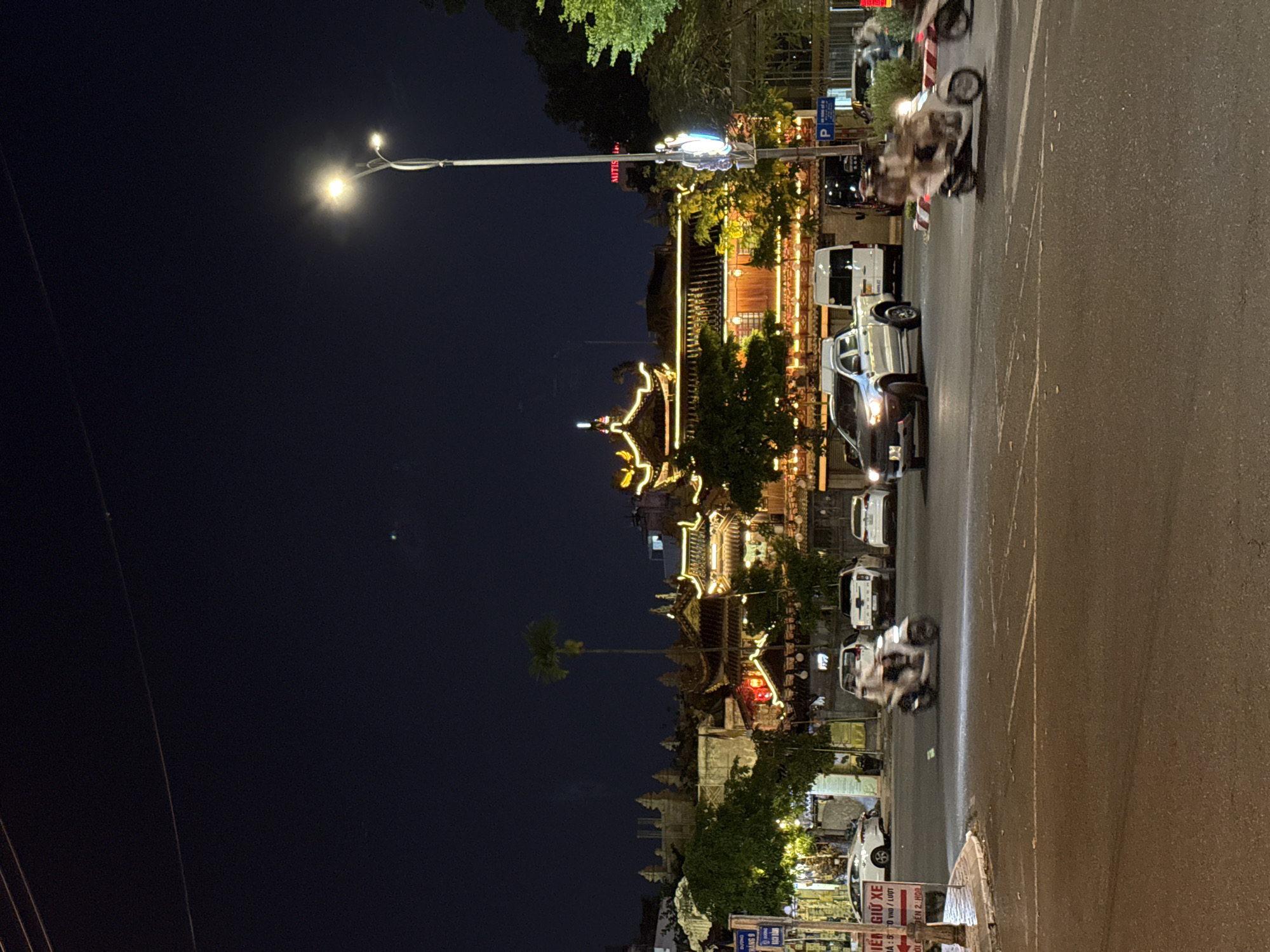 This media file captures a lively city street scene at night in Đà Nẵng, Vietnam. The image is oriented vertically, showing a dark night sky at the top and an asphalt road running horizontally across the bottom and mid-ground.
On the left side of the street, a series of ornate, traditional-style buildings are brightly illuminated with warm string lights and decorative elements, hinting at a cultural or commercial district. Green trees and a tall, slender palm tree stand out against the deep blue-black sky. A prominent streetlight with two bright lamps casts a strong glow near the center-left.
The road features several vehicles, indicating active urban life. A white pickup truck and a white van are visible, along with other cars. Multiple motorcycles are in motion, depicted with motion blur, suggesting typical evening traffic. Some motorcycles appear to be parked or momentarily stopped.
Visible text on various signs provides practical information and local context. A vertical sign displays "MITIS". A standard blue "P" sign indicates parking. Near the bottom right, a sign partially reads "GIỮ XE" (parking, likely motorbike parking), accompanied by "GIÁ: 50 VND / LƯỢT" (price: 50 VND per turn/parking) and "ĐẾN 2 GIỜ" (until 2 AM/PM, likely AM given the night setting). Another sign has the text "HANG 9".
The overall scene conveys the bustling atmosphere of a Vietnamese city after dark, characterized by a mix of traditional architecture, modern vehicles, and extensive artificial lighting.