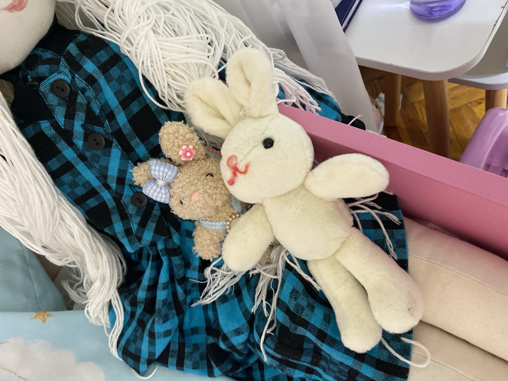 Two stuffed rabbits are positioned on a pink surface in front of a blue and black plaid fabric. The larger, white rabbit is lying on its back with its head facing right. It has black eyes and a red embroidered mouth. A smaller, tan rabbit is positioned above the white rabbit, lying on its stomach with its head to the left. This rabbit has a blue checkered bow on its head and a pink flower on its ear. White yarn is draped around the rabbits and the plaid fabric. The background suggests an indoor setting with wooden beams and white furniture visible. The image appears to have been taken in Lomonosov, Russia.