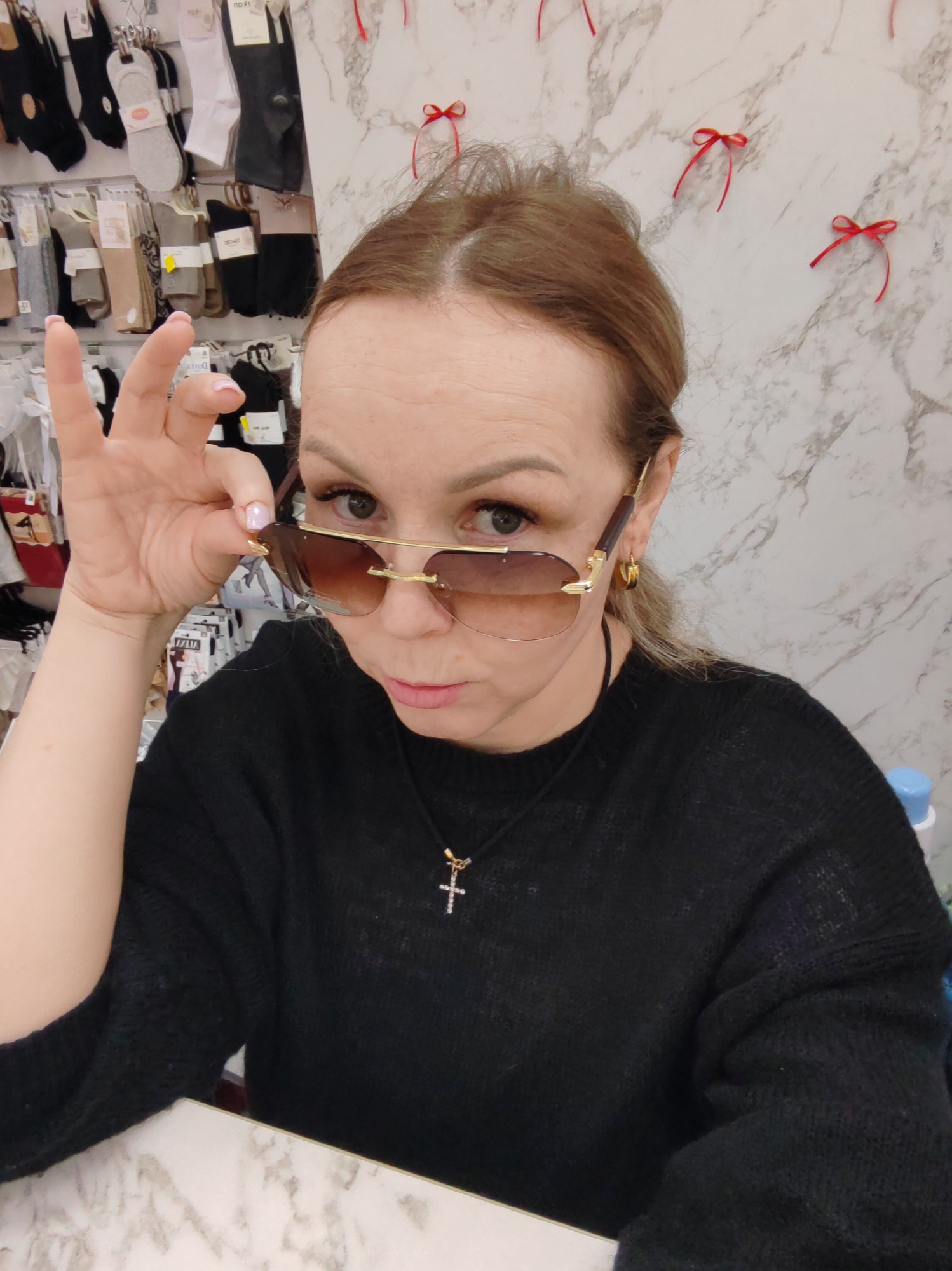 An image captured in a retail establishment in Chaikovsky, Russia, depicts a woman from the chest up, looking directly at the viewer. She possesses light brown hair and light-colored eyes, and wears a black knitted sweater, a black cord necklace with a silver-toned cross pendant, and gold-colored hoop earrings. Her left hand, with light-colored nail polish, holds the right arm of her gold-framed, brown-tinted aviator-style sunglasses, slightly lowering them from her eyes. Her lips are lightly pursed. The background on the left features a display rack laden with numerous pairs of packaged socks in various colors (white, grey, beige, black), indicative of a hosiery section. The background on the right and a surface in the foreground exhibit a white and grey marble-like pattern, with several small red ribbon bows affixed to the wall.