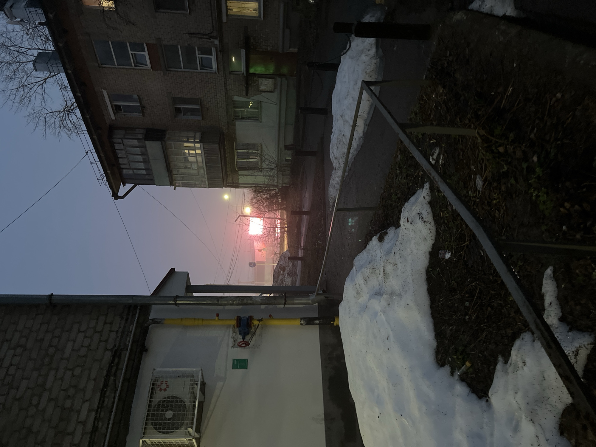 The image shows an urban scene in Chaikovsky, Russia, at dusk or dawn during the winter season. Snow is present on the ground, particularly piled up along a sidewalk edge near a metal railing. Multi-story brick buildings are visible, with some windows illuminated. A gas pipeline is running along a white-painted building, next to an air conditioning unit and a gas valve. In the distance, the setting or rising sun creates a reddish-orange glow on the horizon. The sky is a pale blue-grey. Bare tree branches are silhouetted against the sky near the buildings.