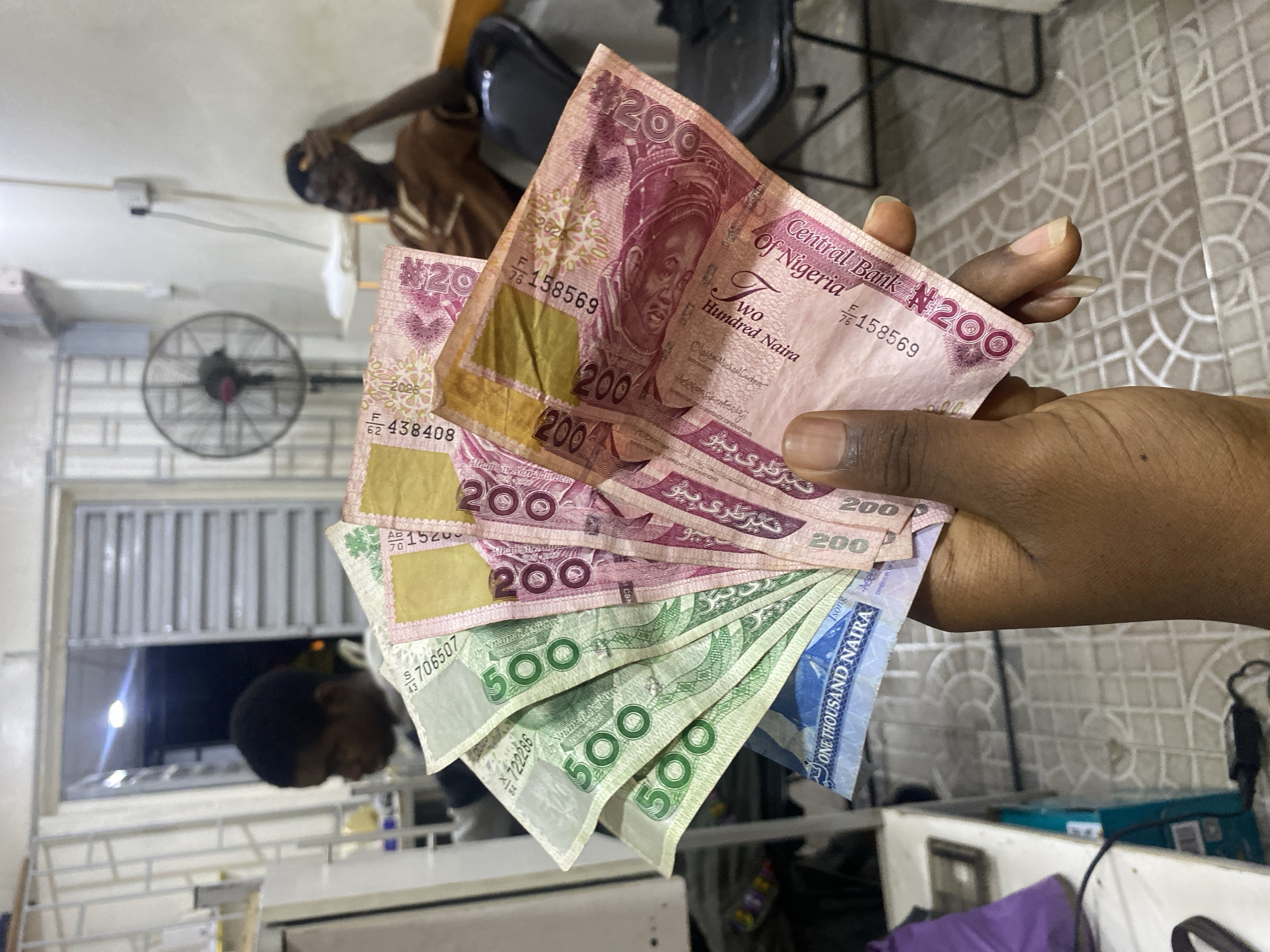 A dark-skinned hand holds a collection of Nigerian Naira banknotes, including several 200 Naira (pink), 500 Naira (green), and one 1000 Naira (blue) note. In the background, two men are visible. One man is lying down, wearing a brown shirt, and the other is in a window or doorway. A fan is mounted on the wall. The location appears to be indoors. It is not possible to confirm that this image was taken in Lagos, Nigeria.