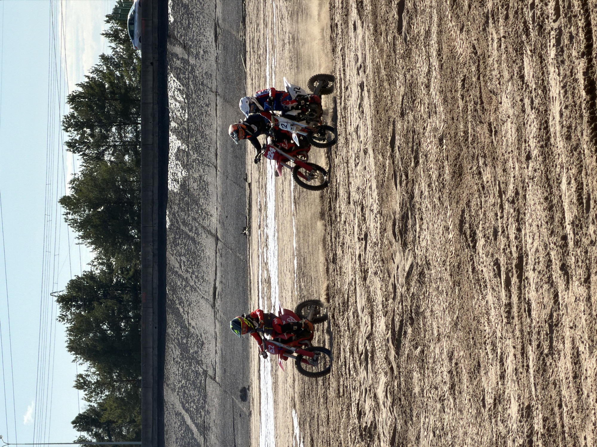 The image depicts a motocross race with two apparent participants in Chaikovsky, Russia. The racers ride on a dirt track bordered by a concrete retaining wall, trees, and power lines. A partial car is visible on the top left. One rider is in the lead, with the number "224" prominently displayed on their motorcycle. Another group of riders are together in the middle. The track appears sandy and dusty.