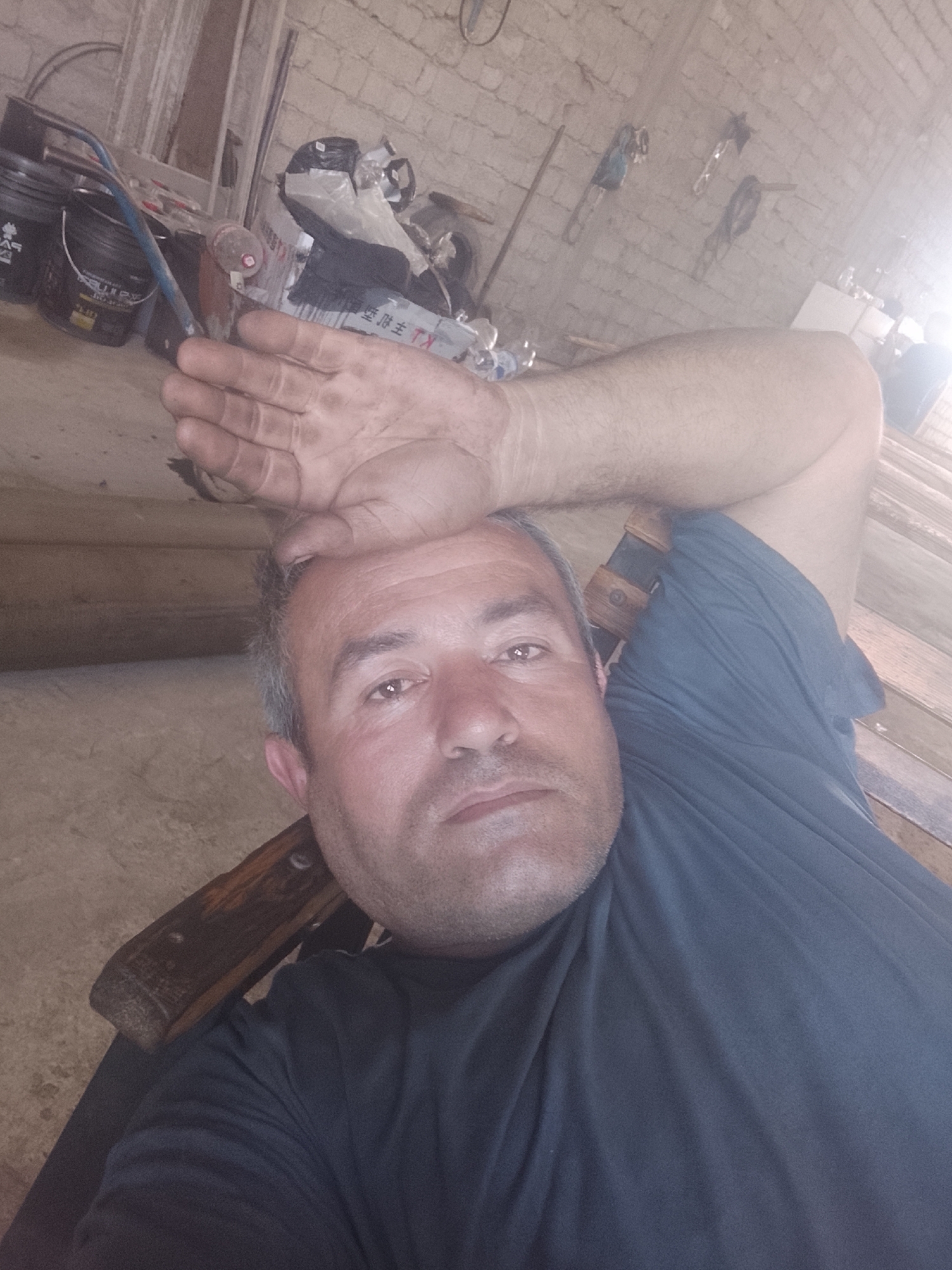 A man is lying down in what appears to be a workshop or storage area, with his hand resting on his forehead. He is wearing a dark blue t-shirt. The background shows a brick wall, various tools and equipment, including buckets and what looks like a wheelbarrow. The lighting suggests it could be daytime, possibly with natural light coming from a window or opening. The overall impression is one of a working environment. The specific location in Sabir Rakhimov, Uzbekistan, is not identifiable from the image itself.