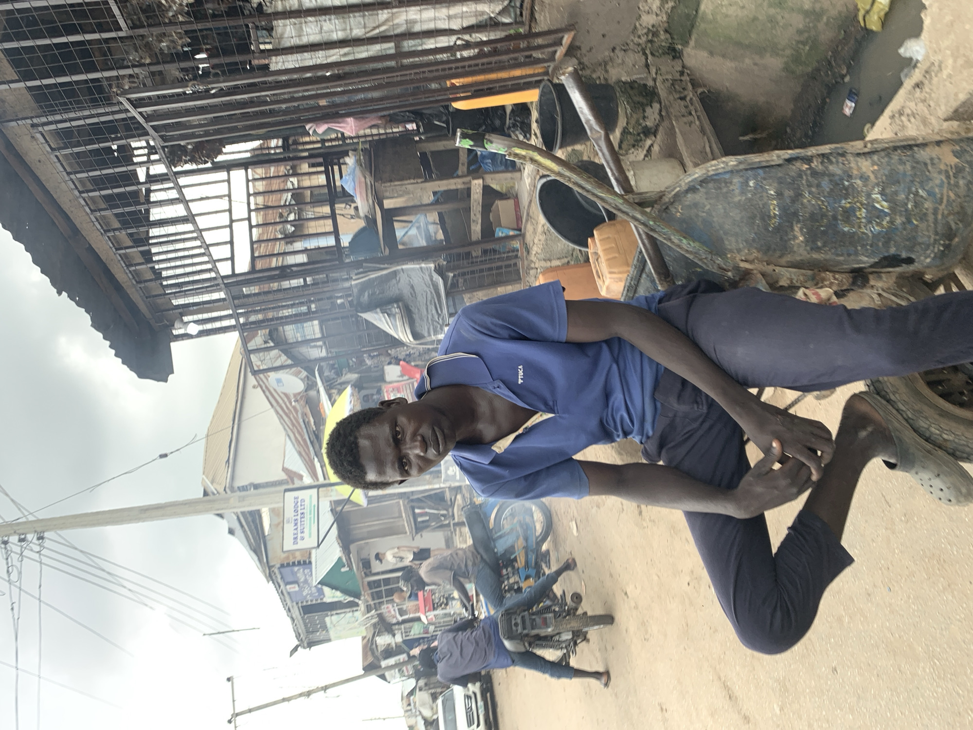 The image shows a busy street scene in Abuja, Nigeria. A young man in a blue polo shirt and dark pants is the main subject, looking directly at the camera. He is leaning on a wheelbarrow, which is partially obscured by his body. In the background, there are several other people, some on motorcycles, suggesting a bustling market or transport hub.
The setting appears to be an outdoor area with various structures, including what looks like market stalls with metal roofing and shelving. There are also signs of disrepair and clutter, with debris on the ground and some overflowing containers. A drainage ditch filled with murky water is visible. The sky is overcast, indicating a cloudy day. The overall impression is of a lively, possibly informal, commercial environment.
One sign in the background reads "DREAM LODGE & SUITES LTD". The clothing of the individuals suggests a warm climate. The general atmosphere seems to be one of everyday activity and commerce.