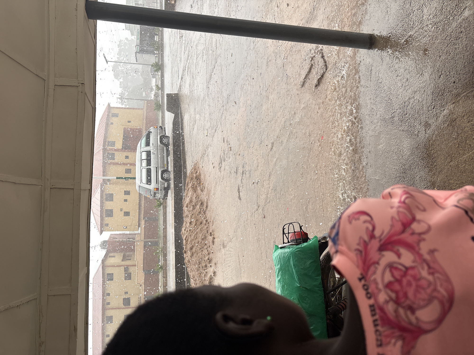 The image depicts a heavy downpour in what appears to be an urban area of Bukuru, Nigeria. The view is from what seems like a covered veranda or window. Rain is sheeting down, creating a blurry, watery effect across the scene. Water is flowing heavily across the pavement.
Visible outside the immediate area is a street, a vehicle, and the facade of several buildings painted in shades of yellow and beige. A dark vertical pole is positioned in the foreground, partially obscuring the view.
In the immediate foreground, there is a dark-skinned person wearing a pink shirt with floral designs and the words "Too Much" on it. A green sack is placed on a bicycle. The person appears to be looking out at the rain.