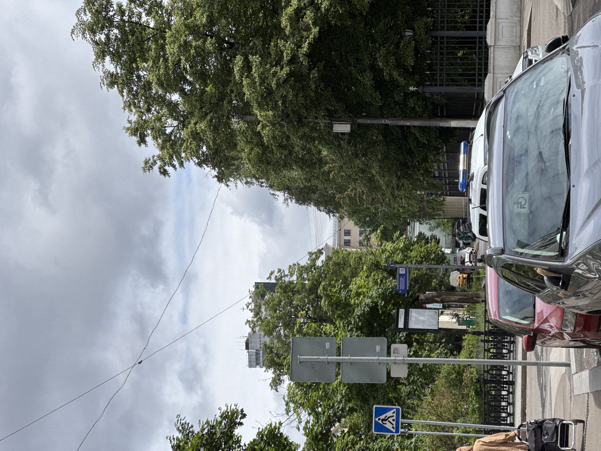 The image depicts a street scene in Moscow, Russia, showing parked cars along a road lined with trees. The sky is cloudy. A pedestrian crossing sign is visible on a pole, along with other street signs. Buildings can be seen in the distance behind the trees. A white van with a blue flasher on top is visible in the distance.