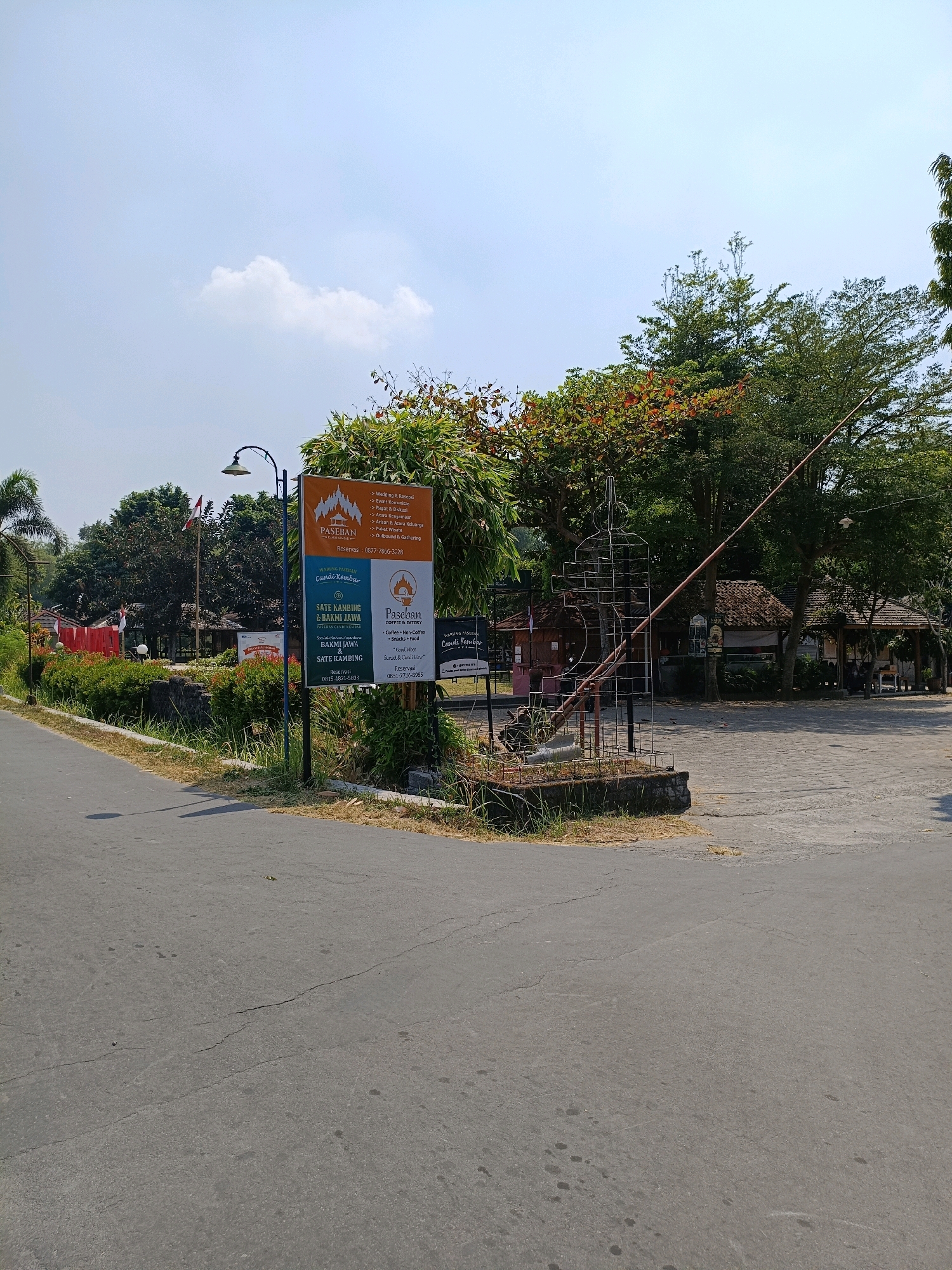 In Bugisan, Indonesia, a roadside view captures a clear, bright sky with a single white cloud. A tall lamp post stands on the left, casting a shadow on the cracked asphalt road. To the right of the road, a large orange and white sign displays information about "PASEBAN Coffee & Eatery" and "WARUNG PASEBAN CANDI KEMBAR". The sign also lists services such as "Wedding & Resepsi," "Event Organizer," and various food items. Behind the sign, lush green trees and some reddish-brown foliage create a dense backdrop. Several small, traditional buildings with thatched roofs are visible further back, nestled amongst the trees. A metal structure, resembling an abstract sculpture, stands in front of these buildings. The overall scene depicts an outdoor area with amenities and natural surroundings.