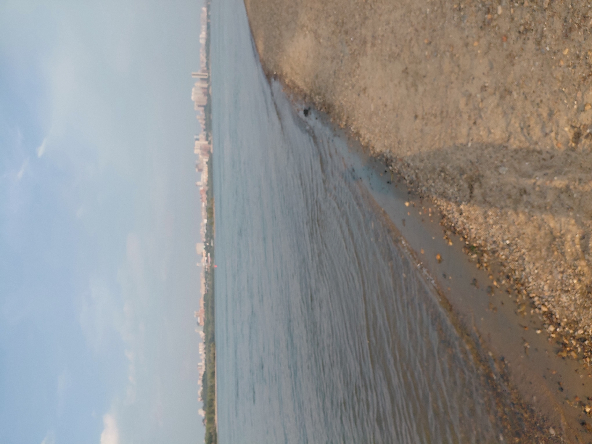The image shows a wide river with a city skyline on the opposite bank. The foreground is a sandy shore with some pebbles and a hint of vegetation. The water appears calm with small ripples. The sky is partly cloudy and pale blue. The city skyline consists of numerous buildings of varying heights, including apartment complexes and possibly some commercial structures. The scene suggests a waterfront area in an urban setting, likely a park or recreational area along the river. Given the context of Novosibirsk, this could be a view of the Ob River embankment.