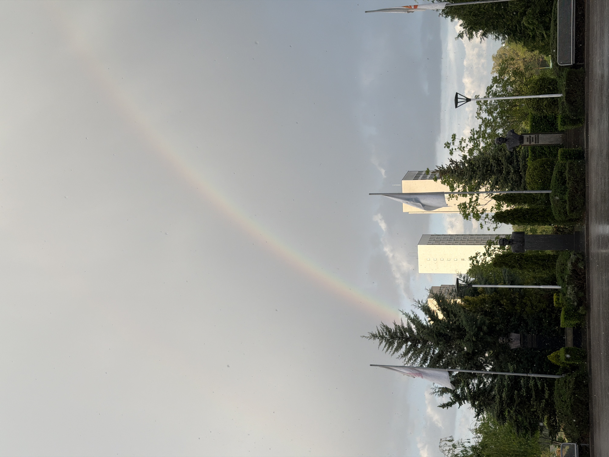 The image shows a rainbow in the sky, ending behind trees near two tall white buildings. In the foreground, there are trees and a statue bust, along with flagpoles. The location is likely Sofia, Bulgaria, given the style of architecture and monuments. It seems to be raining during the picture.