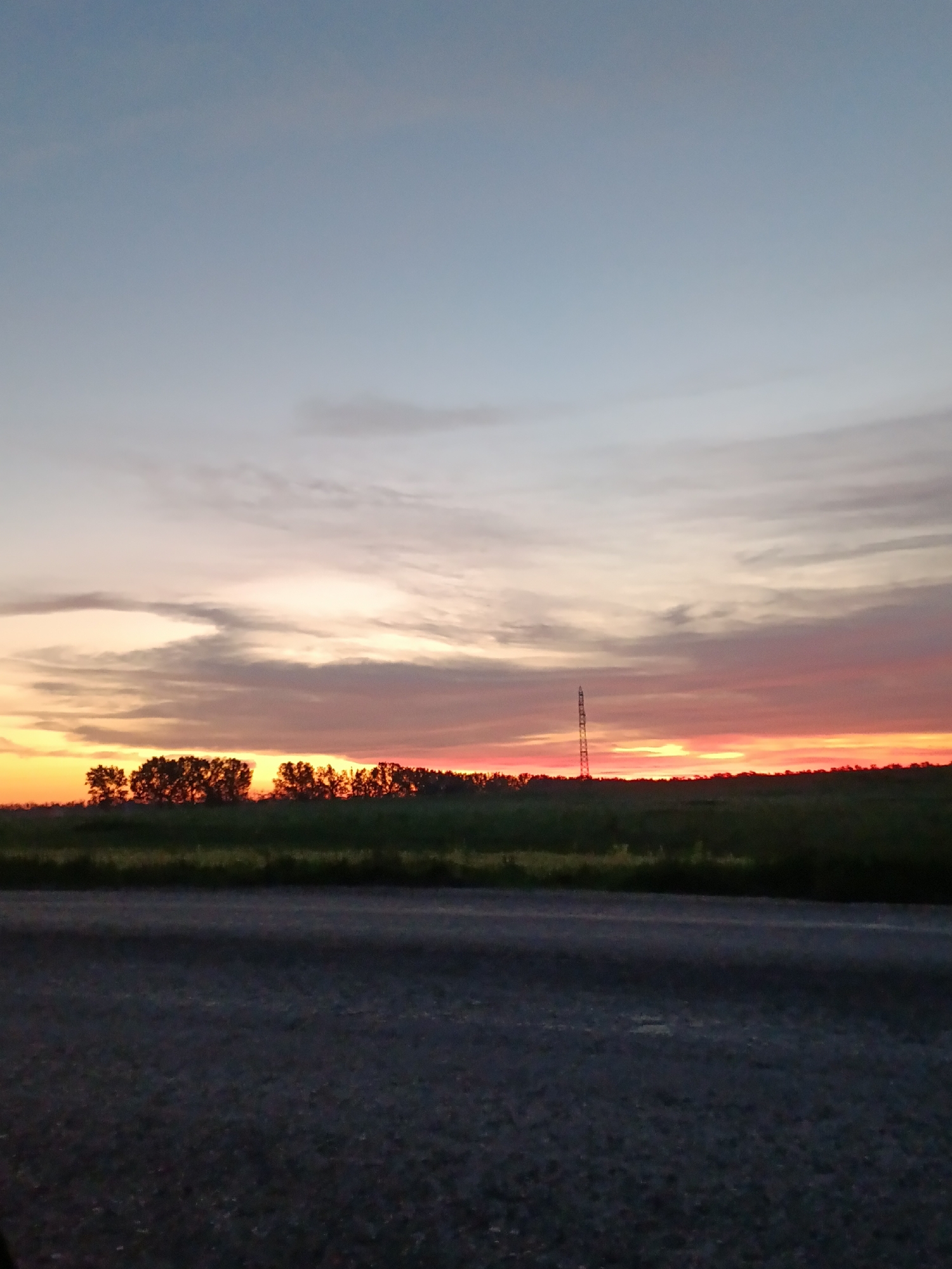 An image depicts a sunset over a rural landscape. The sky exhibits a gradient of colors, transitioning from a pale blue at the top to vibrant oranges and pinks near the horizon. A thin line of trees is silhouetted against the colorful sky. In the distance, a tall, slender structure, possibly a radio tower or power pylon, stands prominently against the sunset. The foreground is dominated by a dark, asphalt road. The scene appears to be set in an open, natural environment.