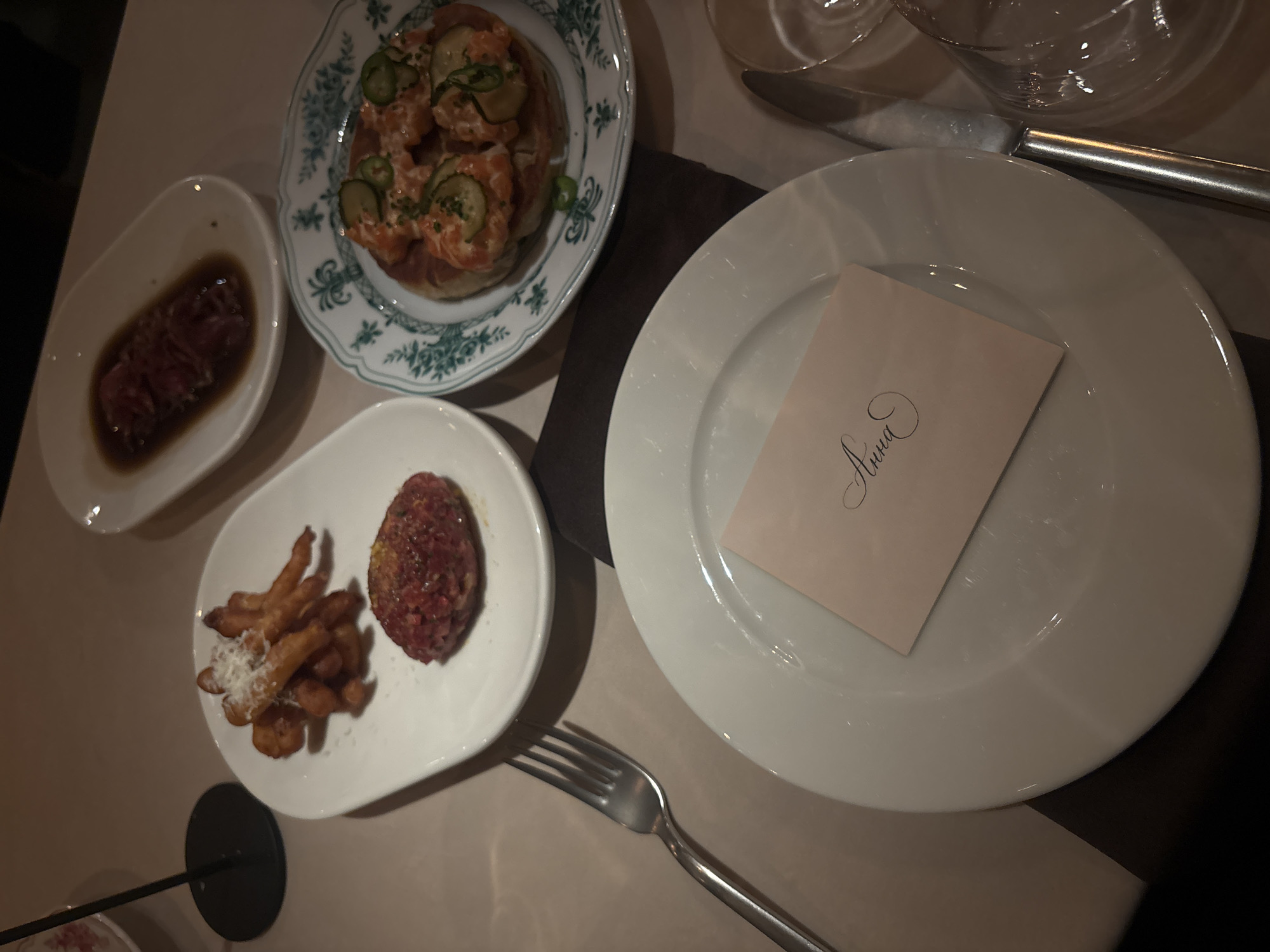 The image captures a dining table setting, likely in a restaurant or private dining room. Various dishes are arranged on the table: one plate contains what appears to be raw beef marinated in sauce, another features a dish resembling shrimp or lobster on toast with cucumber slices, and a third has what seems to be a small mound of minced meat alongside fried potatoes or croquettes. A white plate holds a card with the name "Anna" calligraphically written on it, indicating a reserved seating arrangement. A fork and a knife are also visible on the table. The setting gives the impression of an elegant or upscale dining experience, but it is not possible to tell if it is located in Moscow, Russia.