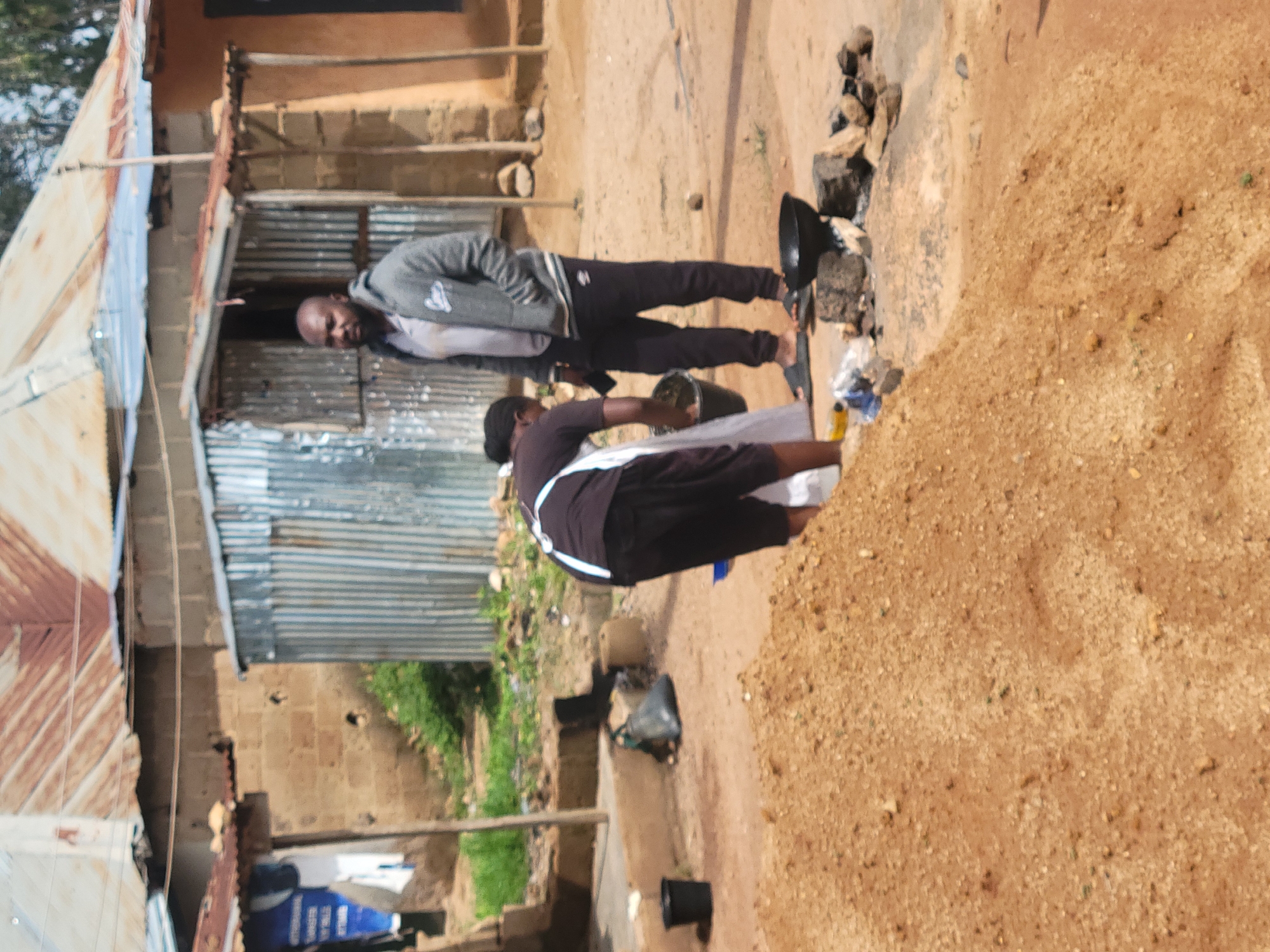 The image depicts an outdoor scene in the city of Jos, Nigeria. In the foreground, a large pile of reddish-brown sand dominates the lower portion of the frame. Behind the sand pile, a person wearing a dark shirt and black shorts, with a white apron tied around their waist, is bent over and appears to be working with a bucket. To the right of this person stands another individual, a man with dark skin and a beard, wearing a gray hooded jacket over a light-colored shirt and dark pants. He is looking towards the person working with the bucket. In the background, there are structures that appear to be dwellings, characterized by corrugated metal roofs and brick or mud walls. Some of these structures have zinc siding. To the far left of the image, a blue sign or poster is visible, partially obscured by a white cloth or garment hanging nearby. There is also evidence of vegetation in the mid-ground. In the mid-ground, a makeshift cooking setup is visible, with stones arranged in a circle around a black cooking pot that is placed over what appears to be embers or a fire. Various discarded items, including a black plastic bag and a black bucket, are scattered on the ground in the mid-ground. The ground surface is predominantly dirt and sand.