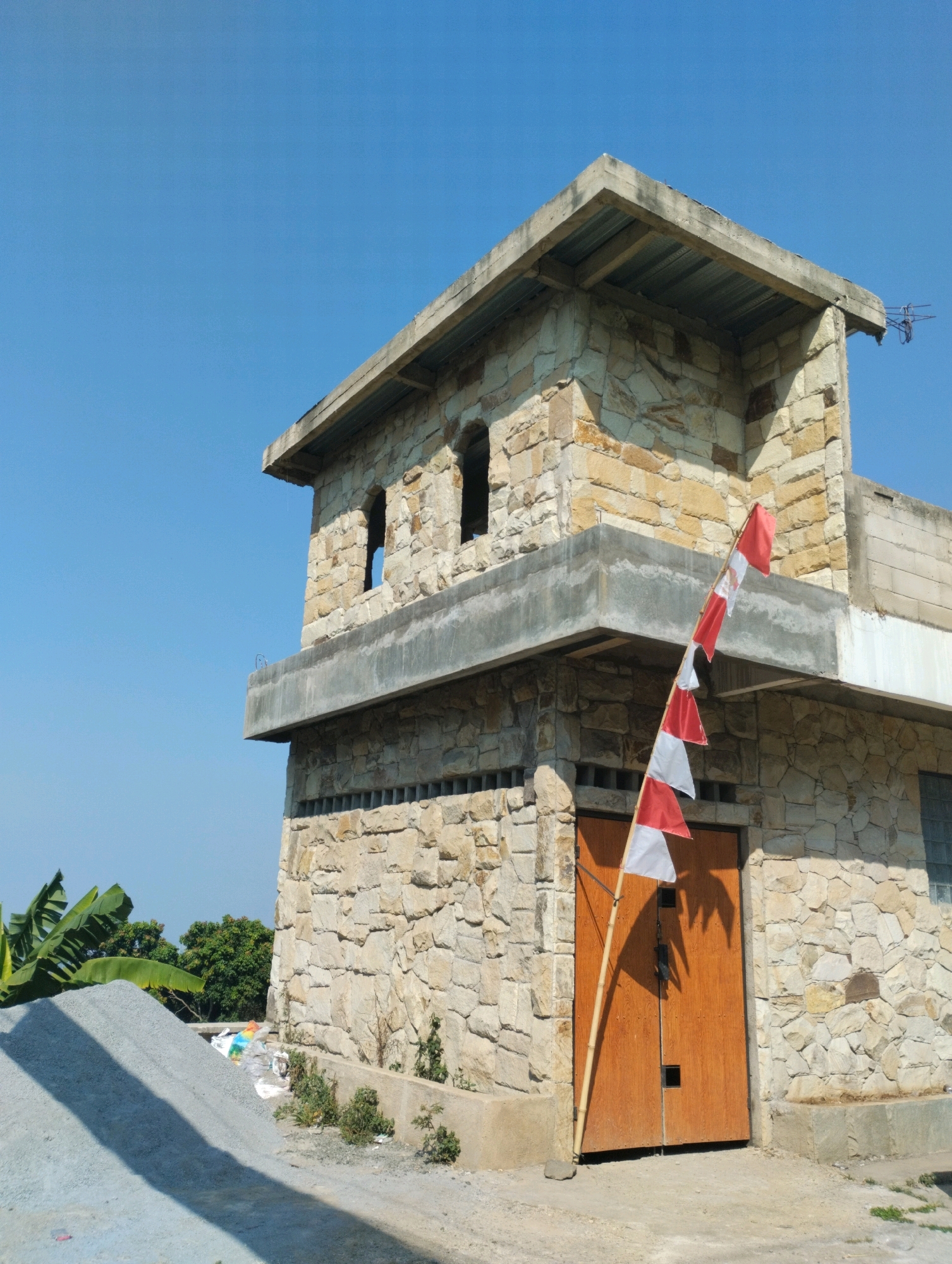 The image depicts an outdoor scene on a sunny day in the city of Cimenyan, Indonesia. In the foreground, a pile of grey aggregate is visible, with banana leaves and trees in the background. Dominating the central and right portions of the frame is a stone building under construction. The building features a multi-level design with rough-hewn, tan-colored stone walls. The upper level has two arched openings. A concrete balcony extends from the middle level. A wooden door is set into the lower part of the building's facade. A bamboo pole with multiple red and white flags, characteristic of the Indonesian flag, is propped against the building near the door. The sky above is clear and blue.