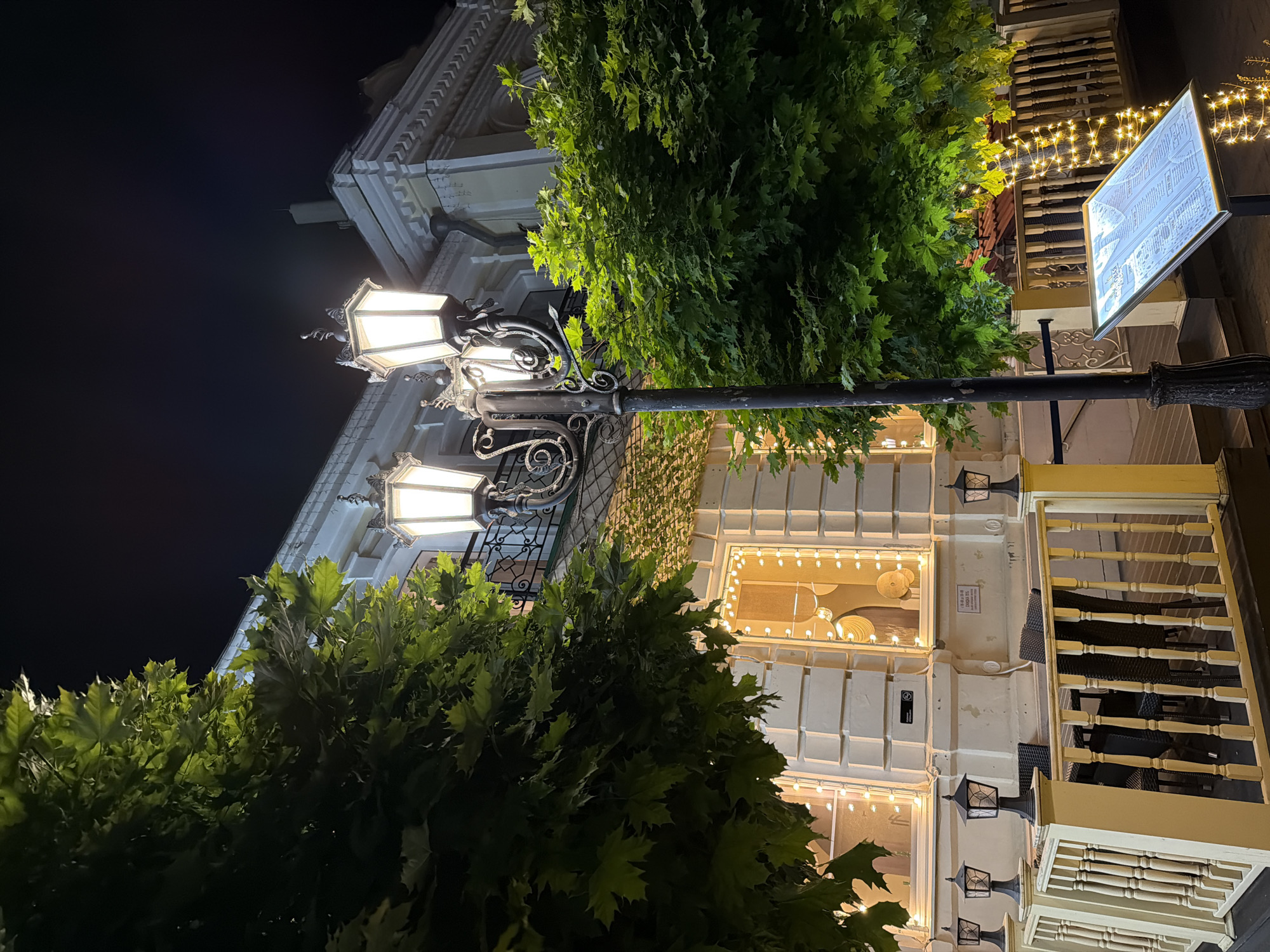 An outdoor seating area of a restaurant or cafe is depicted at night in Kislovodsk, Russia. The area features a wooden railing painted yellow and white, with several small black lanterns mounted on posts. Black wicker chairs and tables are visible beyond the railing. The building in the background is illuminated with warm lights, with a balcony featuring ornate black metal railings. Two large trees with lush green leaves frame the scene, and one tree is wrapped in a string of warm white fairy lights. A tall, ornate street lamp with three illuminated globes stands in the foreground, next to a large, illuminated menu stand displaying a map or directory. The sky is dark, indicating nighttime.