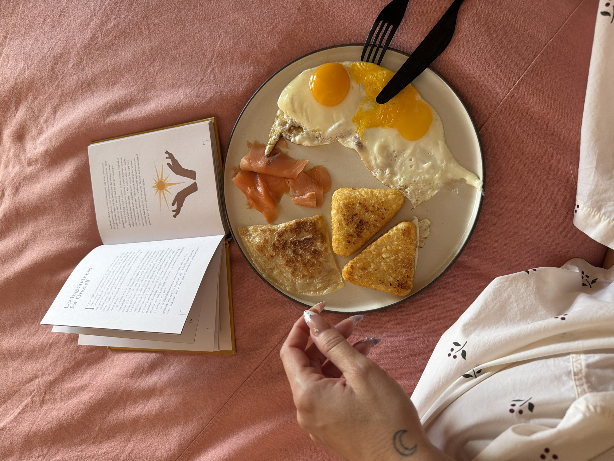 A close-up shot shows a breakfast plate on a pink bed, accompanied by an open book and a person's hand and part of their arm. The plate contains two fried eggs (one with a runny yolk), smoked salmon, and two potato hash browns. Black cutlery (a fork and knife) rest on the upper right side of the plate. To the left is an open book with yellow edges; the visible pages contain text and a simple illustration of hands reaching towards a sun-like symbol. A person's hand, with manicured nails, is reaching towards the plate, while part of their arm, covered in a white and brown patterned pajama shirt, is visible on the right. A crescent moon tattoo can be seen on the person's arm. The background is a pink bedsheet. This image was NOT taken in Mengwi, Indonesia.