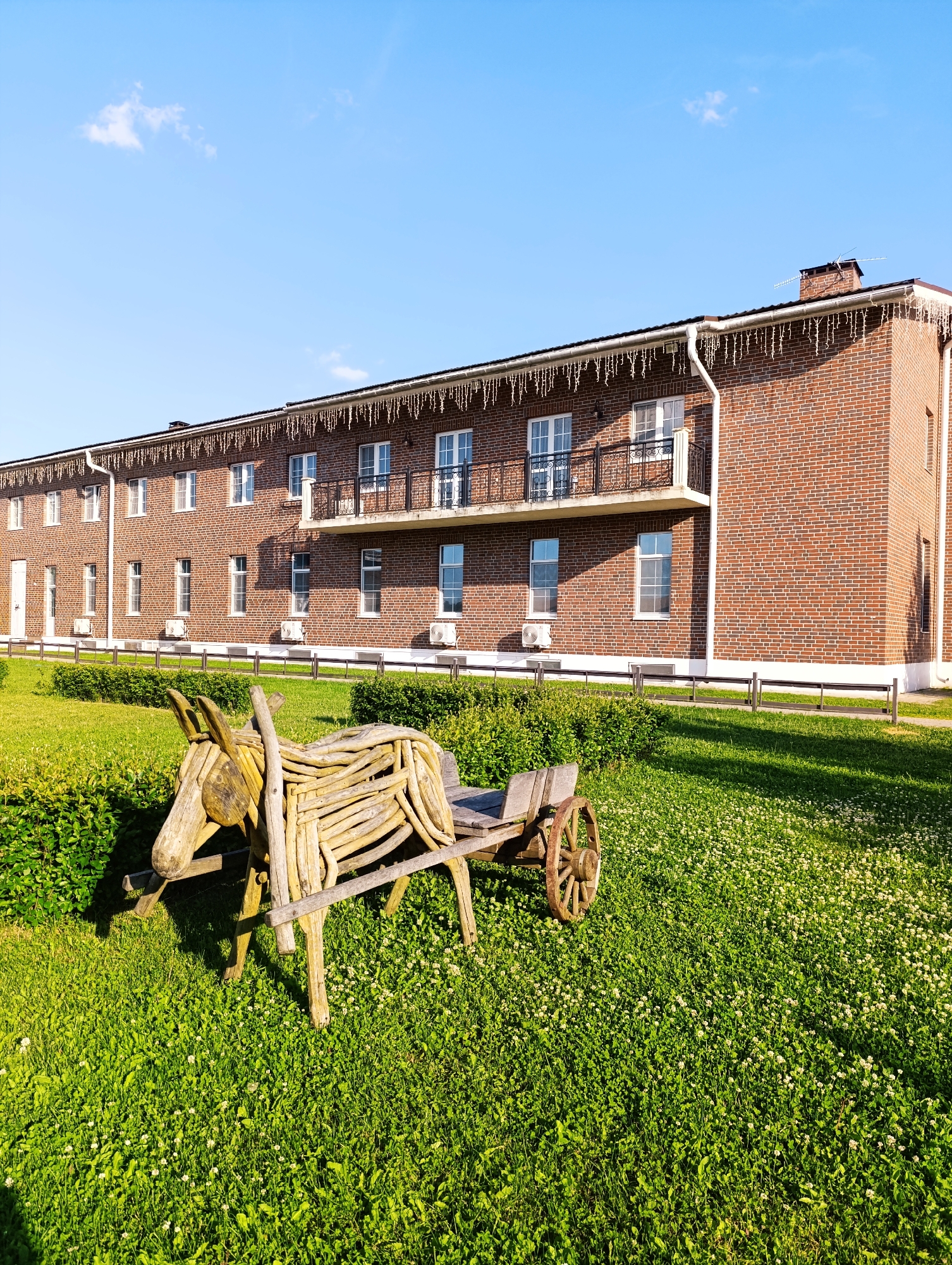 The image shows a rural landscape with a brick building in the background. The building is two stories high with a dark roof and numerous windows. A balcony with a black metal railing is visible on the second floor. White icicle lights are strung along the eaves of the roof. In the foreground, a wooden sculpture of a donkey pulling a cart sits on a green lawn. The donkey is made of light-colored wood, and the cart has a wooden body and a single wooden wheel. The lawn is covered in green grass and small white flowers. A low wooden fence runs along the edge of the lawn in front of the building. The sky above is clear and blue with a few wispy clouds. The scene appears to be a well-maintained outdoor area, possibly a park or a private residence.