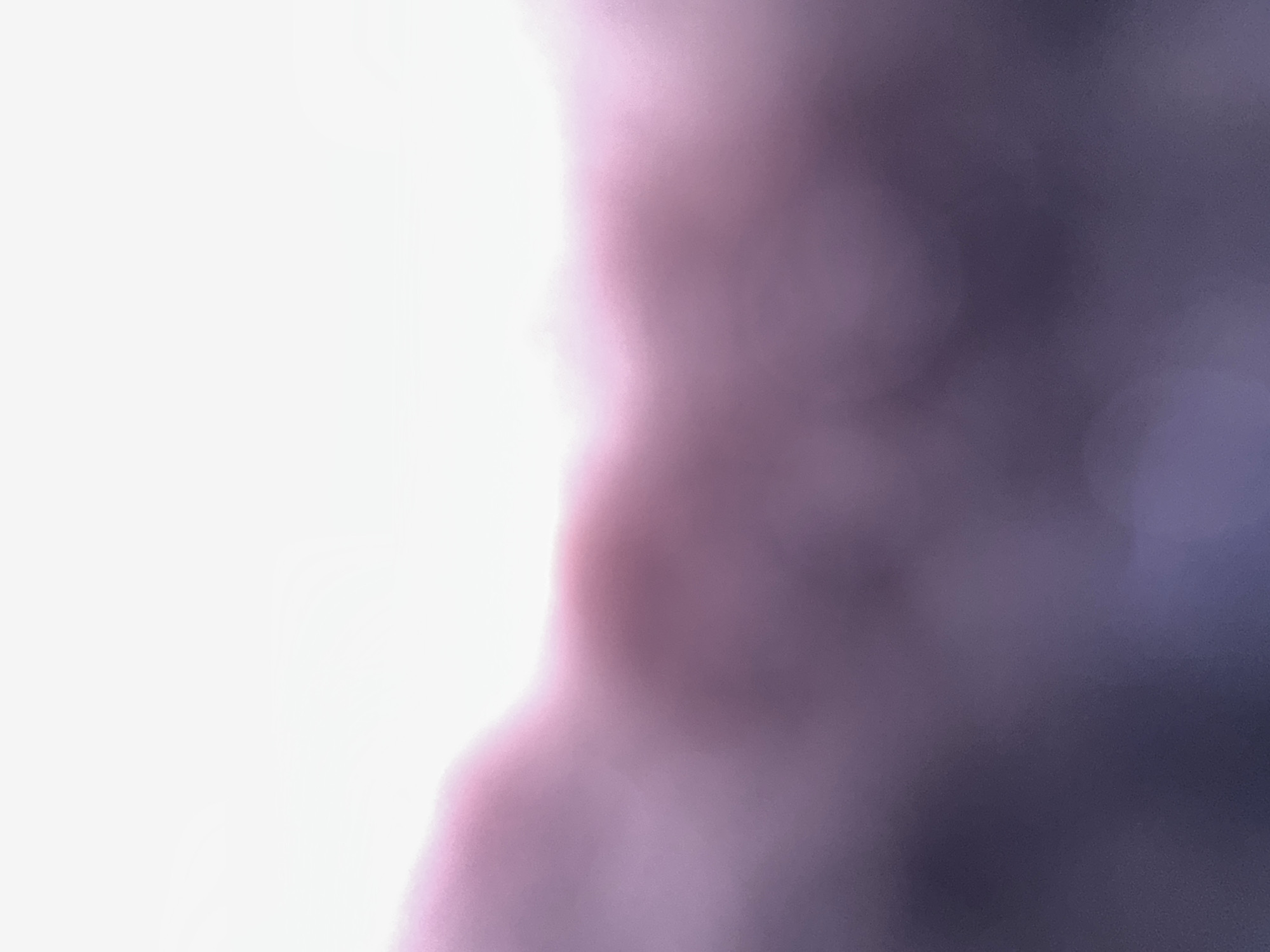 The image is an abstract and out-of-focus depiction of light and color. The left side of the image is a bright, blown-out white, suggesting a strong light source or overexposure. As the image moves to the right, the white transitions into soft, hazy blurs of pink and purple. Further to the right, these colors deepen into a darker, muted purple or deep violet. The overall impression is one of diffused light, possibly from a sunset or sunrise, or a light source filtered through a colored medium or material. There are no discernible subjects, objects, or clear scene elements. No activity or event is visible. There are no location cues, text, or indications of time of day or weather, beyond the suggestion of bright light and atmospheric colors. The lack of sharp focus and distinct forms creates a mood that is ethereal and non-specific.
