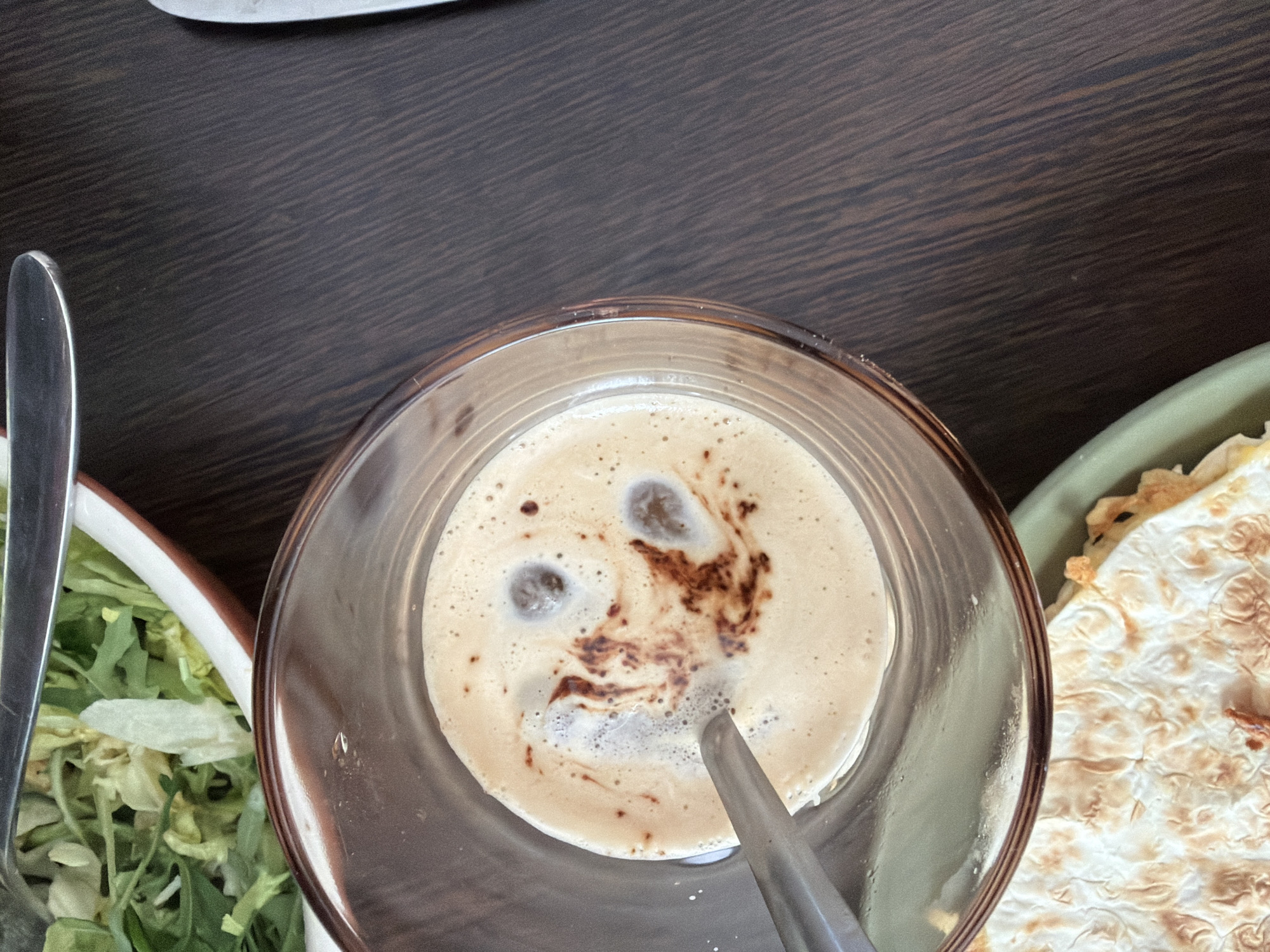 A top-down view shows a glass of light-brown coffee with crema, with dark-brown coffee art forming a face-like pattern. A metal spoon is inserted into the coffee. To the left, part of a salad is visible in a white bowl, and to the right, a piece of flatbread or pastry is on a green plate. The table surface is dark wood. The location is likely a cafe or restaurant setting, possibly in Saint Petersburg, Russia, due to the assumption that the user requesting information about this image is located in Saint Petersburg, Russia. No people are visible, and the focus is on the food and drink arrangement.