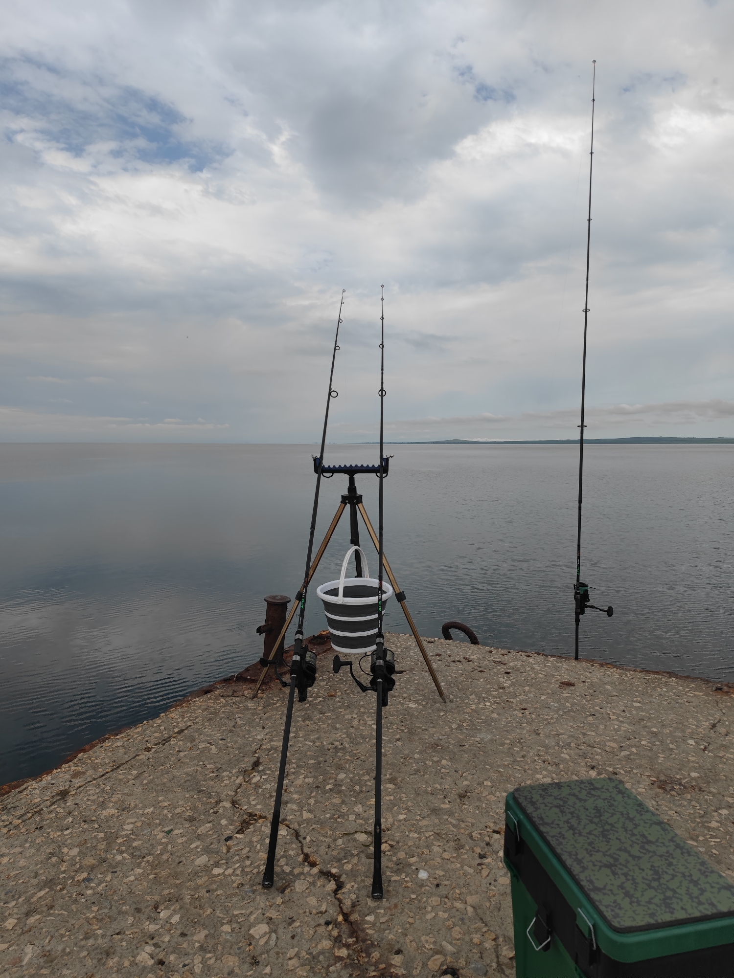 The image shows a fishing scene on a concrete pier. Three fishing rods are set up, two in a tripod stand with a collapsible bucket hanging below, and one standing alone. A green fishing tackle box is on the pier to the right. The background is a calm body of water under a cloudy sky. The pier is old and shows signs of wear. It may be in Sennoy, Russia.