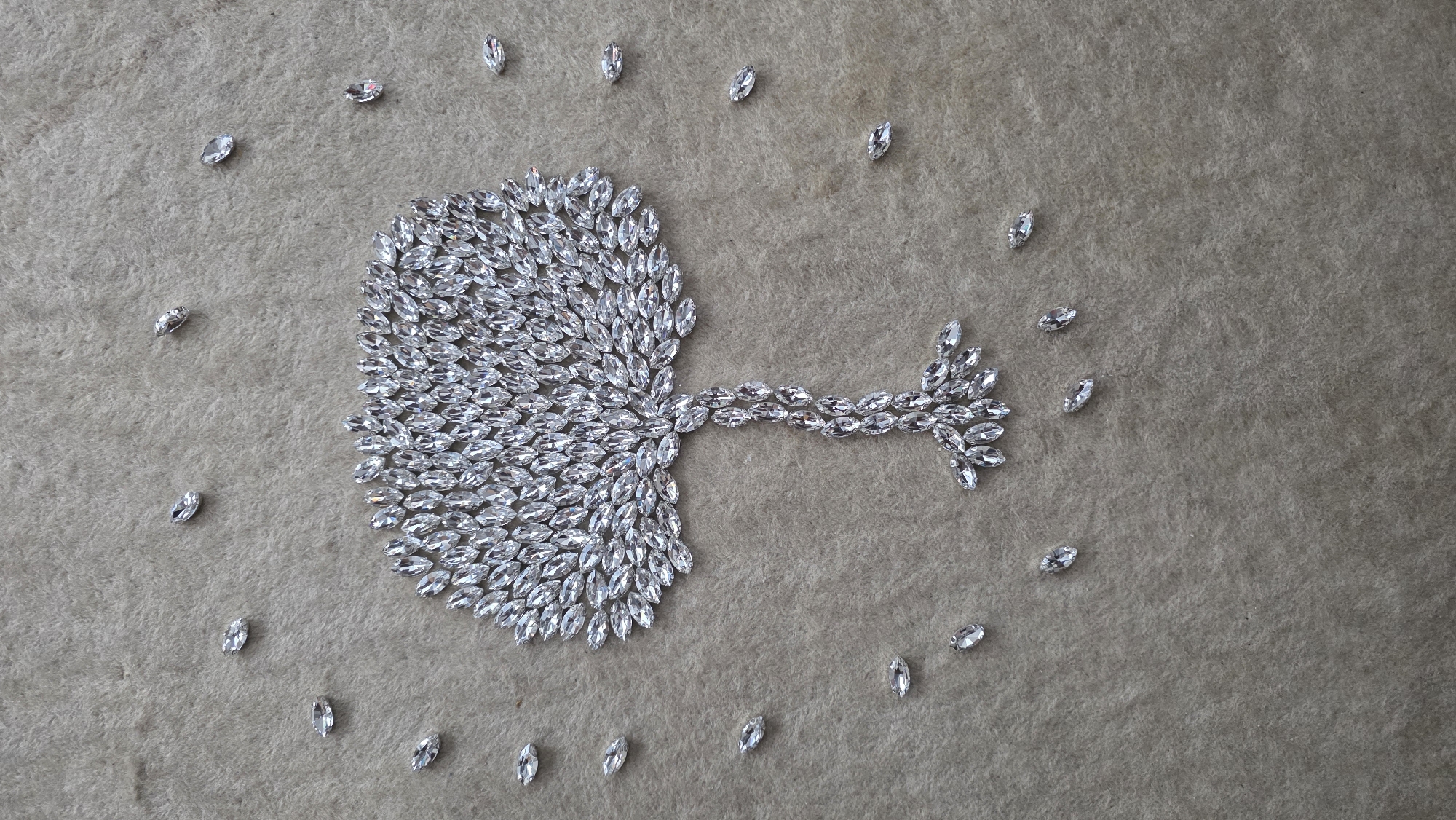 The image presents an artistic arrangement of numerous clear, marquise-cut rhinestones or crystals on a soft, textured, light brown or beige surface, likely felt or carpet. The primary subject is a central, abstract shape composed of many closely packed rhinestones, resembling a stylized tree or an organic form like a lung, with a broad, dense section on the left transitioning into a narrower, branching stem-like structure extending to the right. The facets of the rhinestones glint under even lighting, creating a shimmering effect.
Around this central arrangement, individual marquise-cut rhinestones are scattered in a loose, somewhat circular pattern, suggesting a frame or additional decorative elements. No people are present, and no specific activity is taking place; the image captures a static, decorative display. The background is uniform and lacks distinct features that would indicate a specific time of day or outdoor weather conditions. The setting appears to be an indoor surface. There is no visible text in the image. While the location context is Blagoevgrad, Bulgaria, there are no visual cues within the image itself to confirm this.