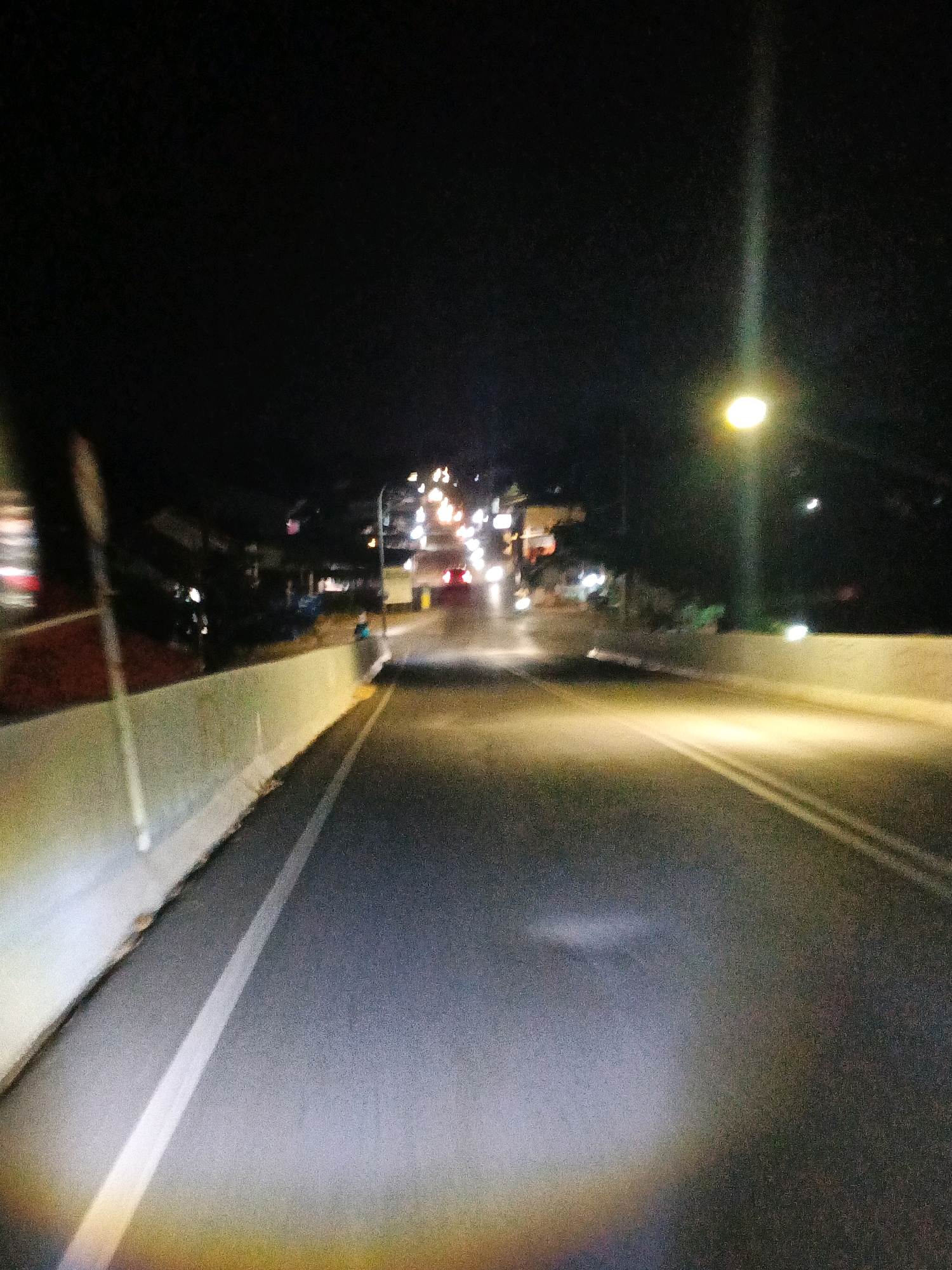 The image depicts a nighttime scene on a road in Sirau, Indonesia. A streetlamp illuminates the roadway and the surrounding area. The road appears to be a two-lane highway with a white dividing line. There are buildings and lights visible on the side of the road in the distance, suggesting a town or village. The lighting conditions are low, with the primary illumination coming from the artificial light sources. The image has a slightly blurred quality, possibly due to motion blur or low light conditions. There are no people clearly visible in the image, nor any specific actions or interactions taking place. The environment appears to be an urban or semi-urban setting at night.