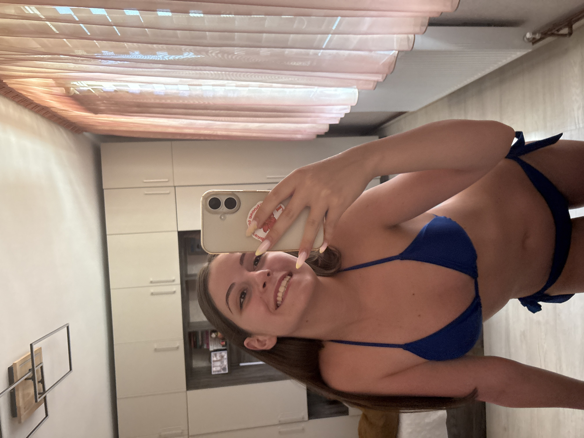 A young woman is taking a selfie in a bedroom. She is wearing a blue bikini and smiling. The room has white cabinets, a ceiling light fixture, and pink curtains. The floor is made of light-colored wood.