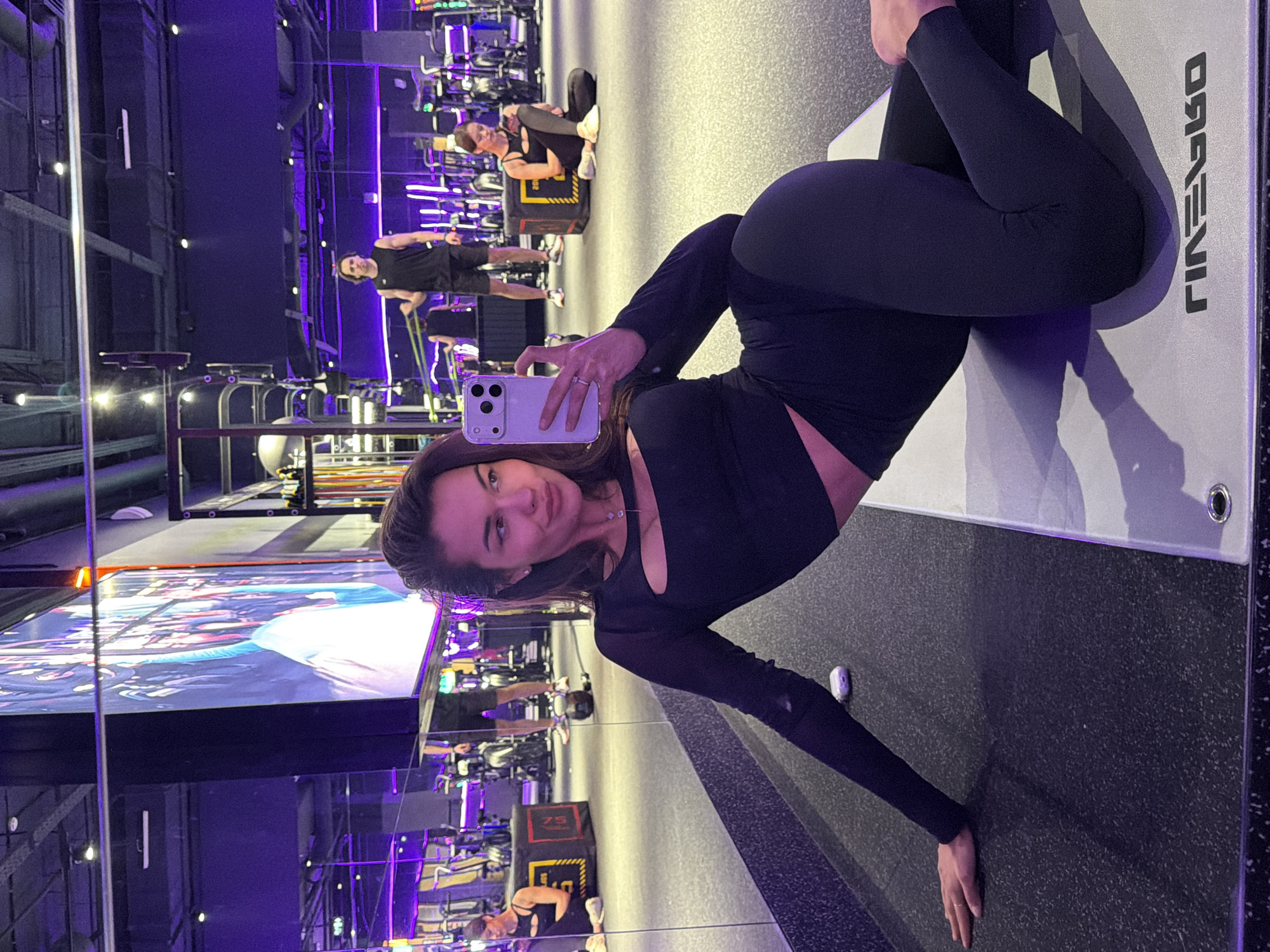 The image shows a woman taking a selfie in a gym. She is wearing black workout clothes and is positioned on a gray workout mat. In the background, other gym-goers are visible using exercise equipment. The gym has a modern design with purple lighting and mirrored walls. Based on information available online, the gym is likely located in Krasnoyarsk, Russia.