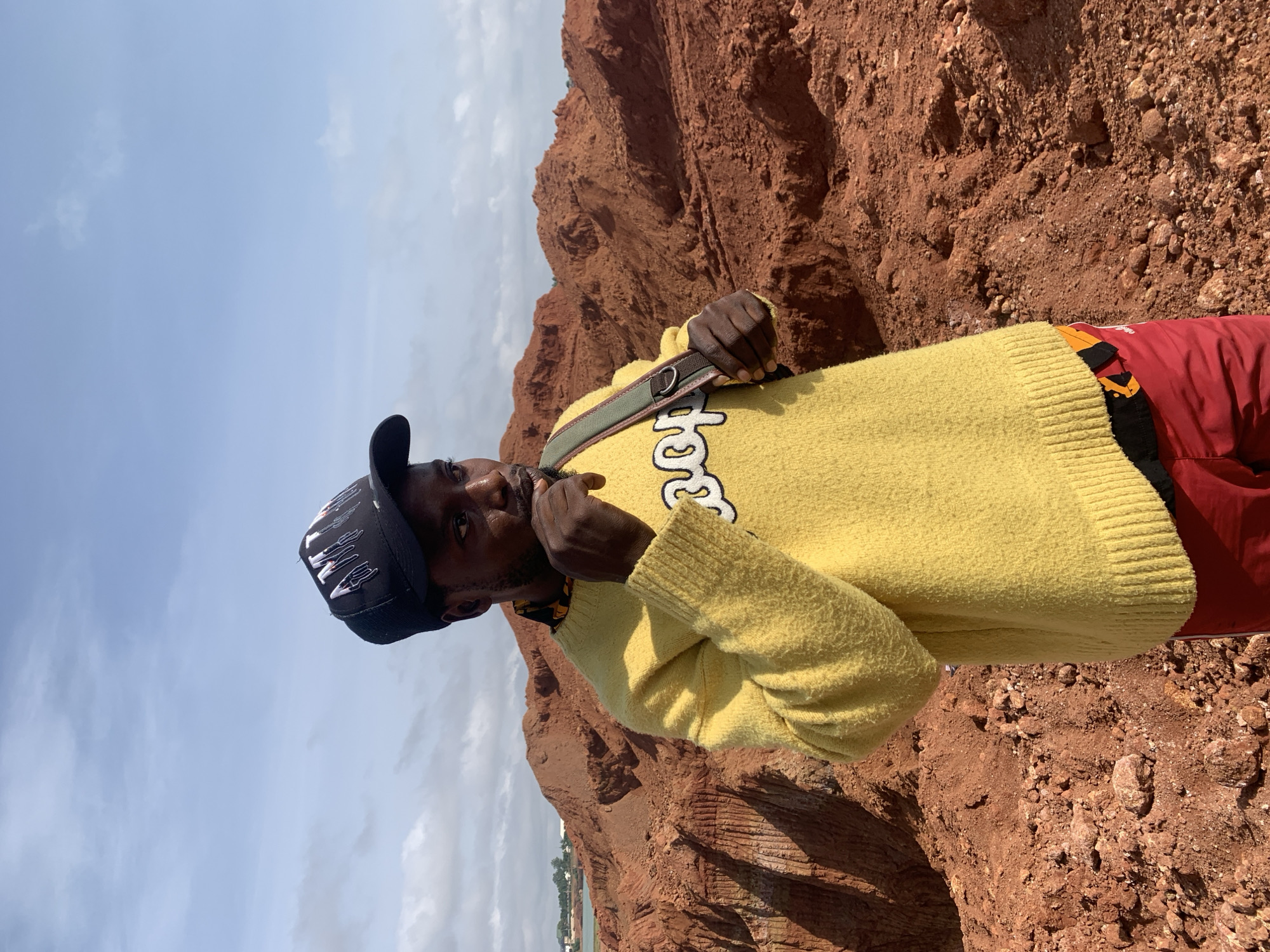 A young Black man wearing a yellow sweater, a black baseball cap with white and red graphics, and red shorts stands in a quarry. The background consists of mounds of red earth and a cloudy sky. The man is looking off to the right, with his right hand touching his mouth. A strap from a bag is visible over his right shoulder. The terrain appears to be a pit or excavation site.