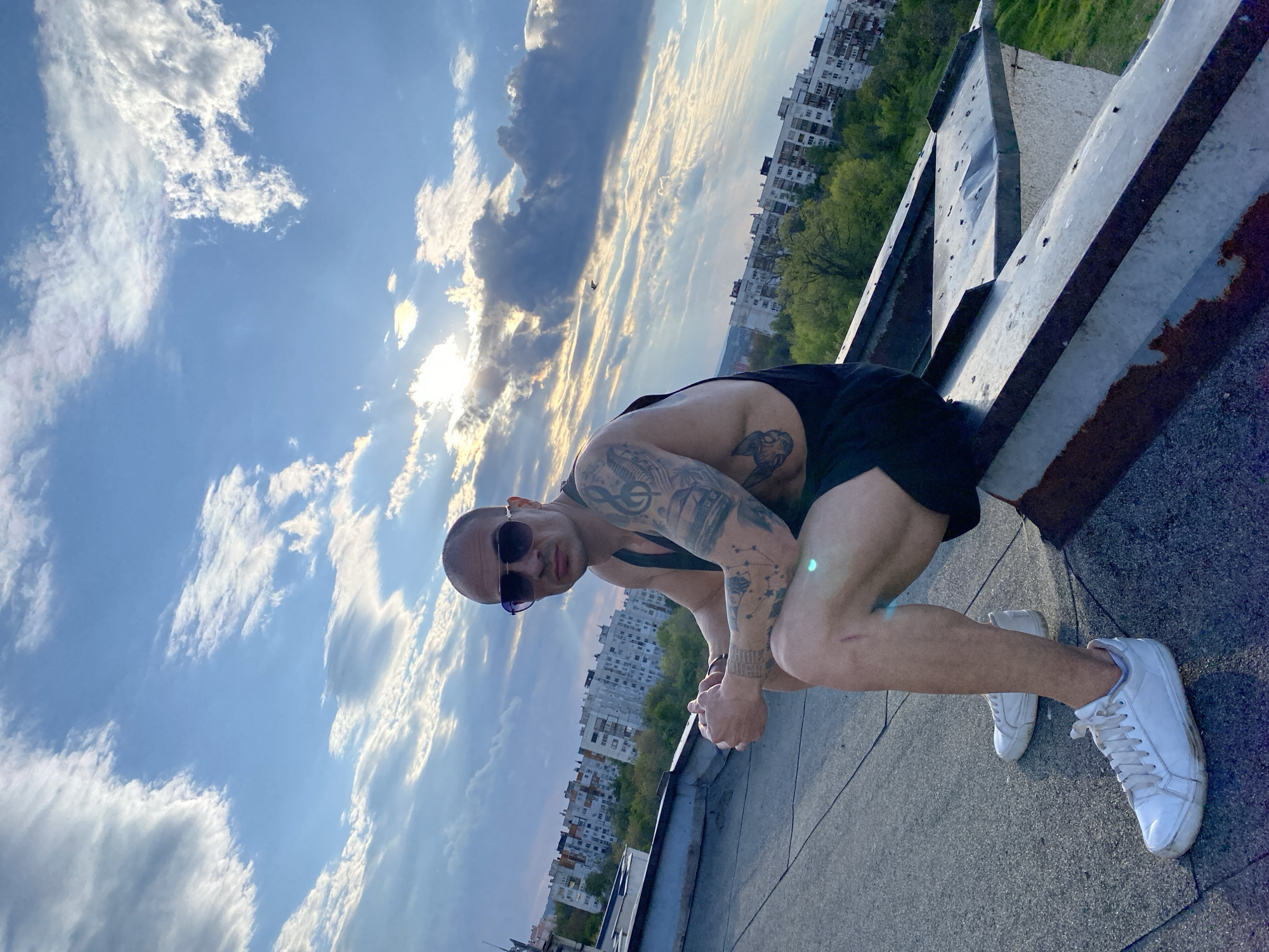A bald man with dark sunglasses, wearing a black tank top, black shorts, and white sneakers, is captured in a crouched pose on a rooftop. He looks directly at the camera, with a neutral expression. His arms are extensively covered in tattoos, and specific text "YOUNG JUICE" is visible on his left forearm.
The setting is a rooftop in Pleven, Bulgaria, overlooking an urban landscape. In the background, numerous multi-story apartment buildings with repeating window patterns are visible, interspersed with green trees. The sky is bright blue with scattered white clouds, and the sun is prominent, creating a strong lens flare and long shadows, suggesting either late afternoon or early evening. The rooftop itself is composed of concrete or similar material, with some weathered metal structures along the edge.
