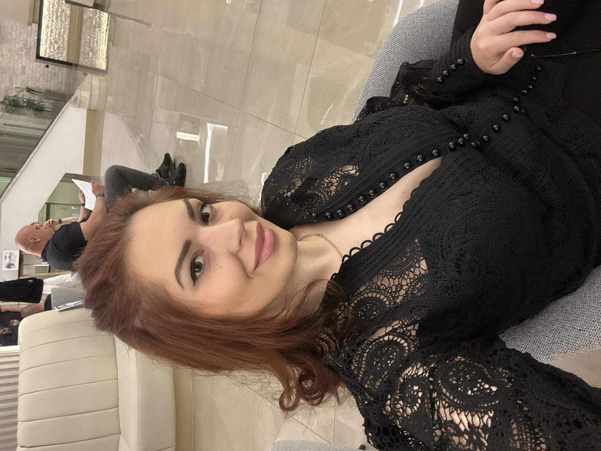 The image shows a selfie of a woman with auburn hair, wearing a black lace top with decorative buttons, posing indoors. In the background, out of focus, a man is sitting and reading. The setting appears to be a modern interior with light-colored tiled flooring. The location of Kazanlak, Bulgaria, cannot be determined from the provided image.