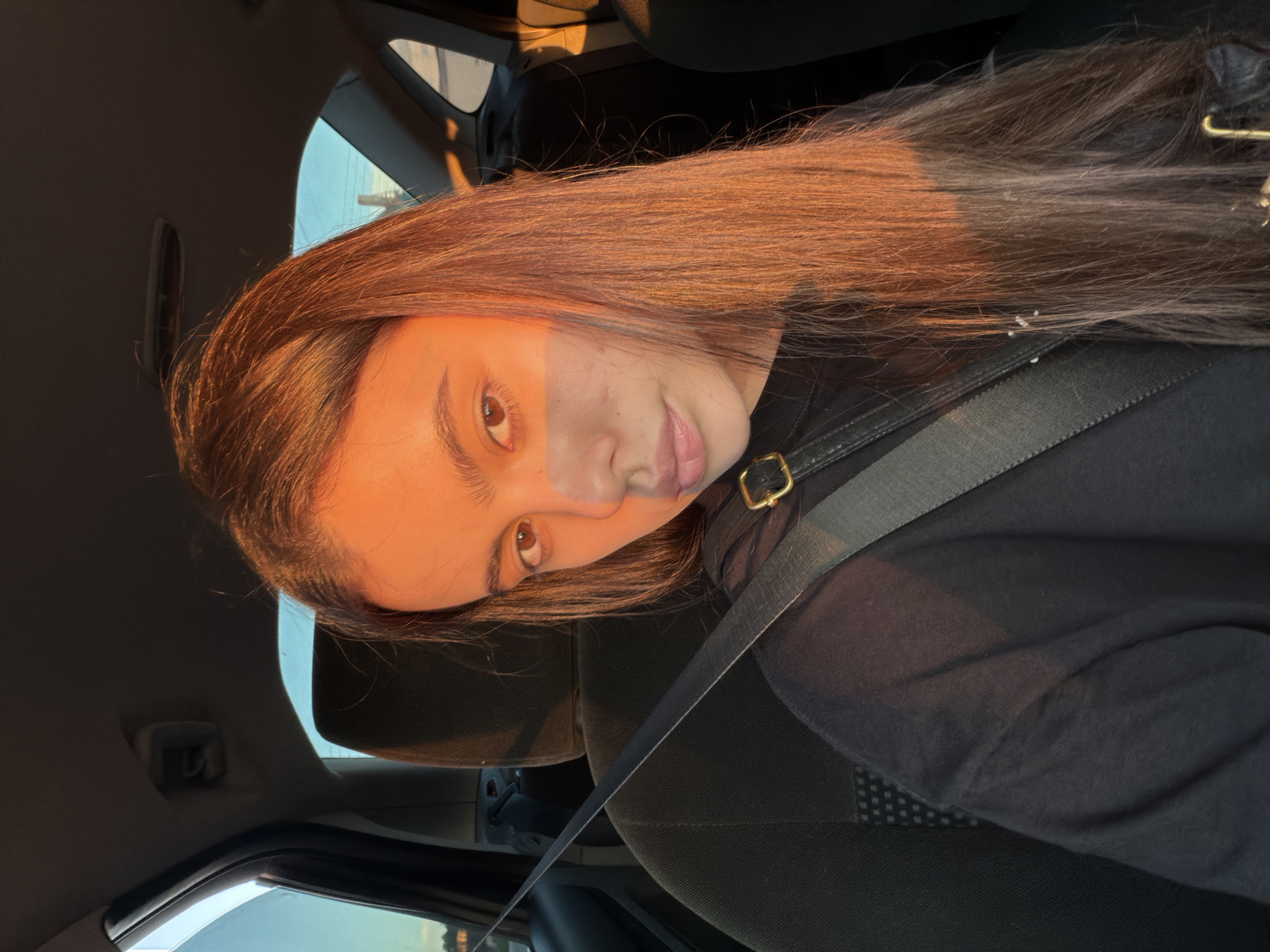 A young woman with long, dark hair is seated in the passenger seat of a car. The sun is setting, casting a warm, orange glow on her face and hair. She is wearing a black t-shirt and her seatbelt is fastened. The interior of the car is visible, including the headrest of the driver's seat and the window. The scene appears to be in Chaikovsky, Russia, judging by the style of the car's interior and the surrounding scenery visible through the window.