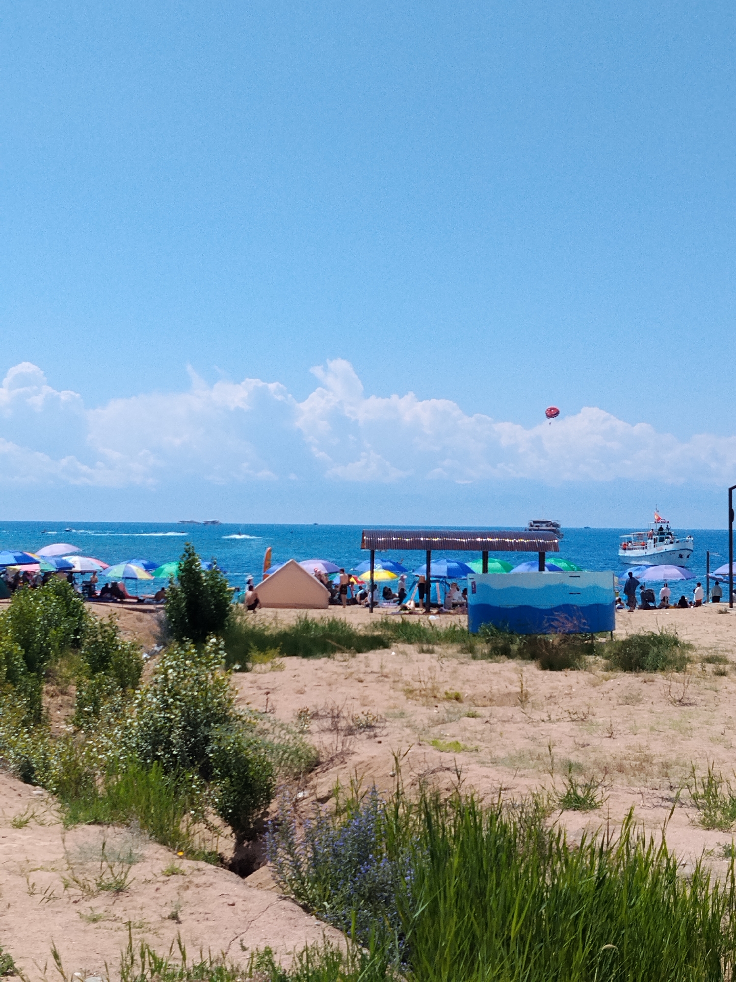 This is a wide-angle, eye-level, outdoor, daytime, full-shot photograph of a crowded beach on a sunny day with a clear blue sky and scattered white clouds. The foreground is dominated by a sandy beach with sparse, dry vegetation and some taller green grass in the lower right corner.
In the middle ground, a beach scene unfolds with numerous beach umbrellas in various colors, including blue, green, pink, and white, providing shade for beachgoers. Several people are visible sitting or standing under the umbrellas and on the sand. A small, light-colored tent or shelter is set up on the beach, and a blue and white structure, possibly a changing room or a play area, is present.
The background features the blue ocean with several boats and jet skis on the water. A boat is seen to the right, and a paraglider is visible in the sky above the water. The horizon is marked by the distant shoreline and a line of trees. The overall atmosphere is that of a lively recreational area.