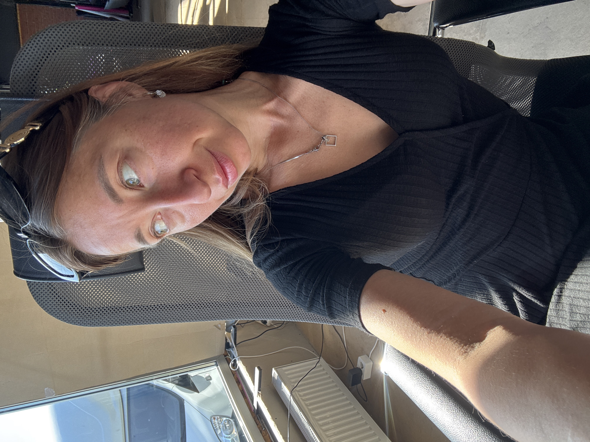 A Caucasian woman with light brown hair and green eyes is seated in a black office chair. She is wearing a black, ribbed, V-neck top and a silver necklace with a geometric pendant. Sunglasses are perched on her head. The photo appears to be a selfie taken at a tilted angle. In the background, a portion of a white radiator, wiring, and a window with a view of a silver vehicle can be seen. The location is indicated as Chaikovsky, Russia.