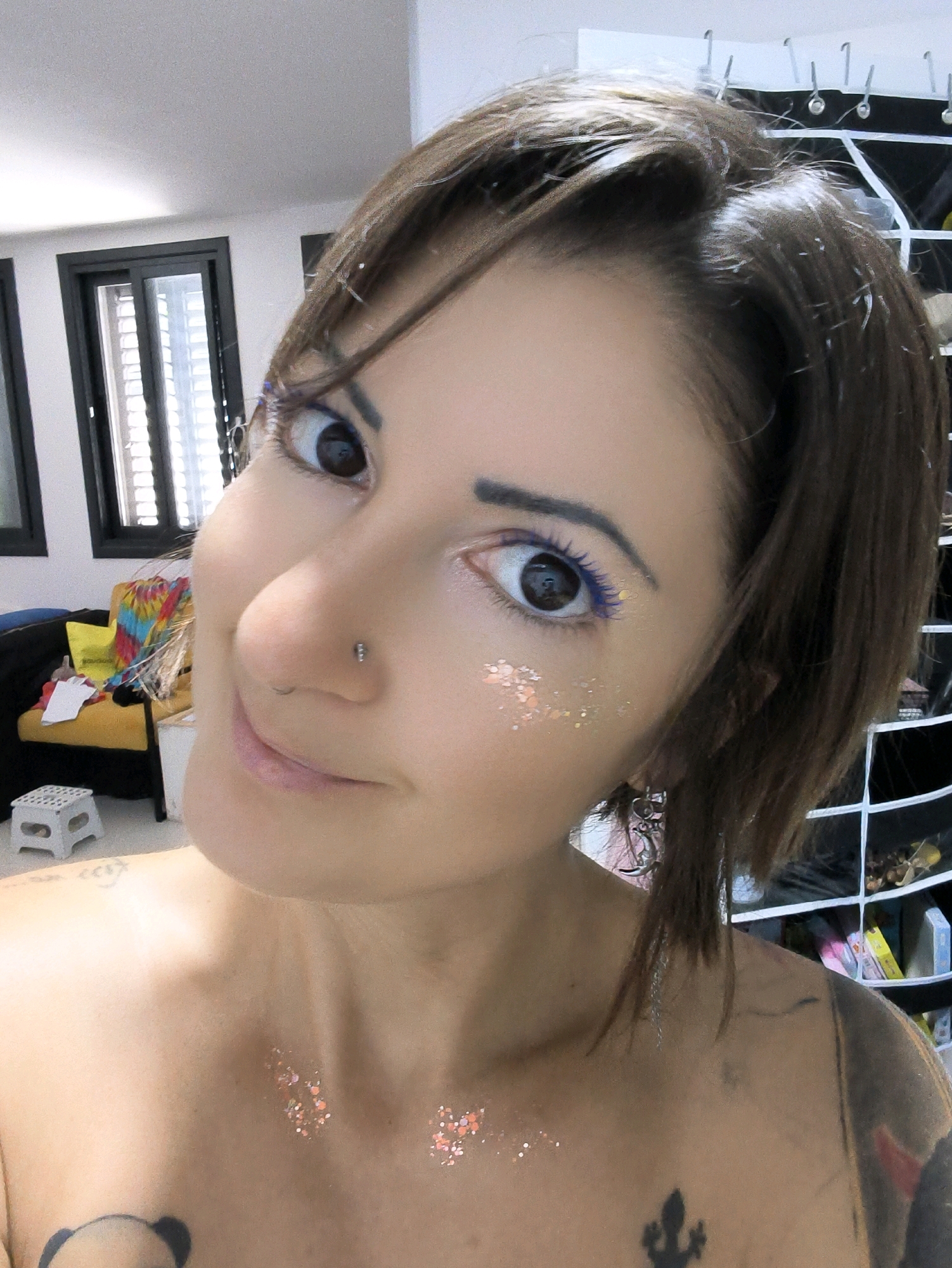 A close-up, shoulder-level, medium shot shows a young woman with short brown hair and a nose piercing. She is wearing blue mascara and has glitter applied to her cheeks and collarbone. Her left eye is in focus, revealing a brown iris. She is facing the camera with a slight smile. In the background, a yellow chair and a white stool are visible. A wall with shelves holding various items can be seen to the right. The environment appears to be an indoor setting, possibly a room in a house or an apartment. The image was taken in the city of Lachish Regional Council, Israel.
