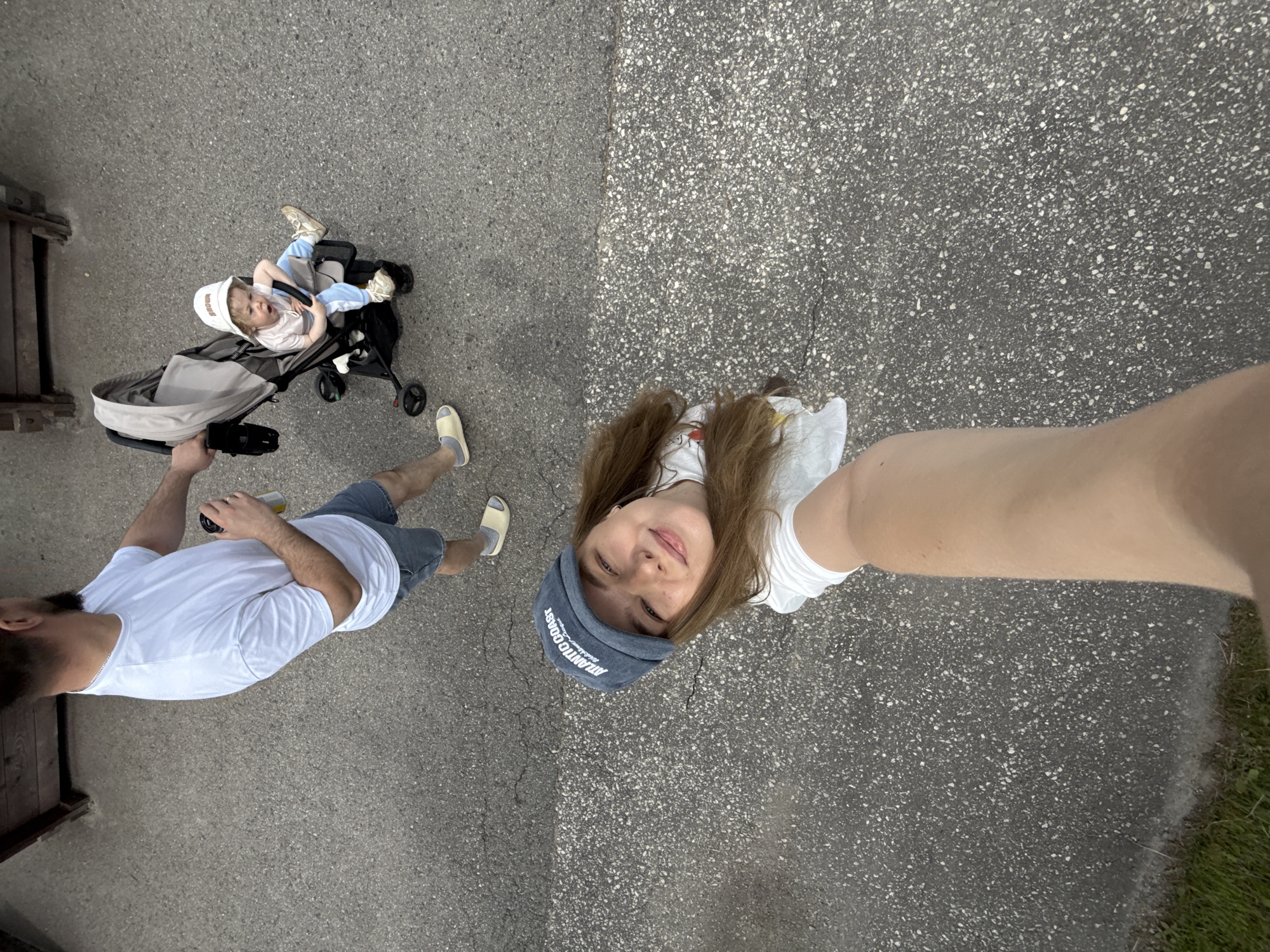 This high-angle selfie captures three individuals – a woman, a man, and a baby – during an outdoor walk in Dobrograd, Russia. The camera is held by the woman, whose arm and smiling face are visible on the right, looking up towards the lens. She wears a white t-shirt and a dark blue cap with the text "ATLANTIC COAST" partially visible. Her long, brown hair falls over her shoulders.
To her left, a man is seen from the chest down, wearing a white t-shirt, denim shorts, and light-colored sandals. He holds a metallic beverage can in his right hand and pushes a dark grey baby stroller with his left. In the stroller, a baby is seated, wearing a light-colored hat (with illegible text), a light pink top, and light blue pants with white shoes. The baby's mouth is open, looking towards the left.
The setting is an outdoor paved area, likely a path or sidewalk, with two distinct ground textures: a darker, speckled asphalt and a lighter, smoother concrete, meeting at a clear seam. A strip of green grass is visible along the far right edge of the frame, and a dark wooden structure or fence is partially seen on the far left. The scene takes place during the daytime under soft, possibly overcast, lighting, suggesting a casual family outing.